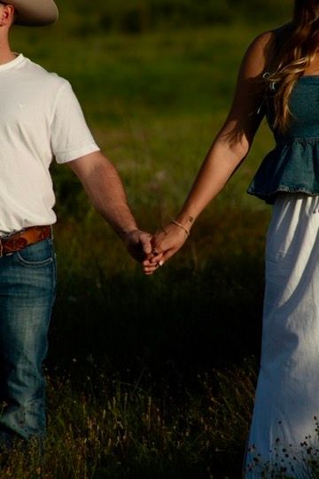 A couple holding hands outside in a field at sunset, wearing casual white and blue outfits.