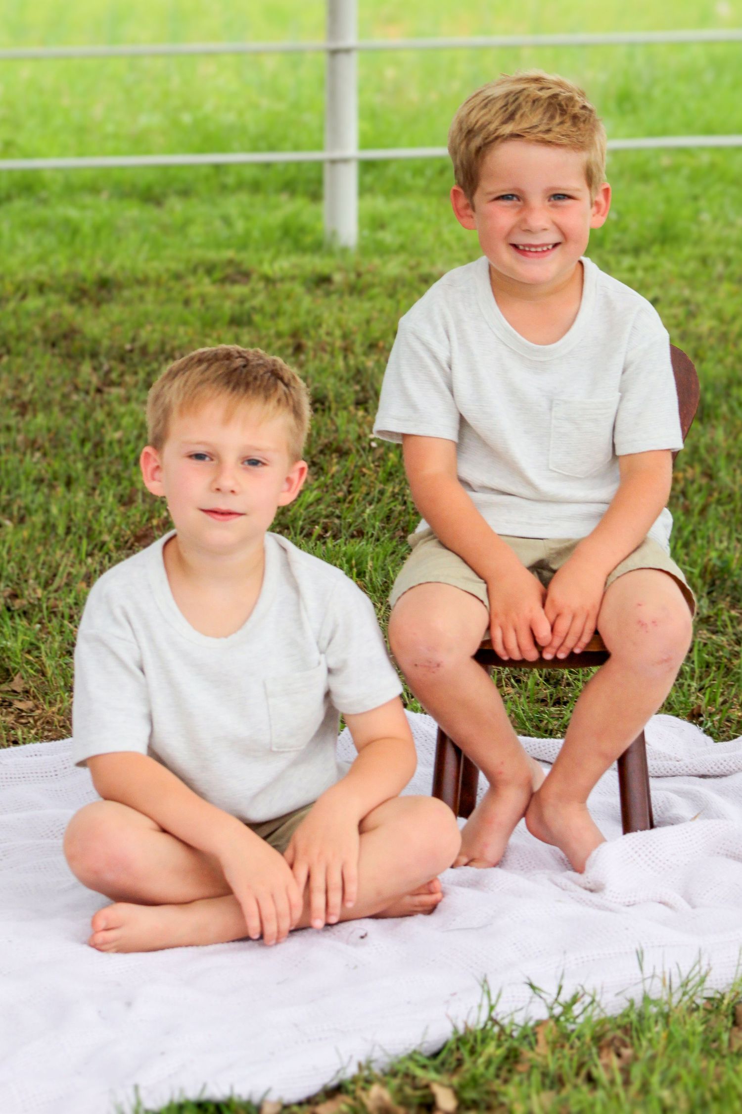 Two boys in matching white shirts sitting on blanket outdoors for casual portrait.