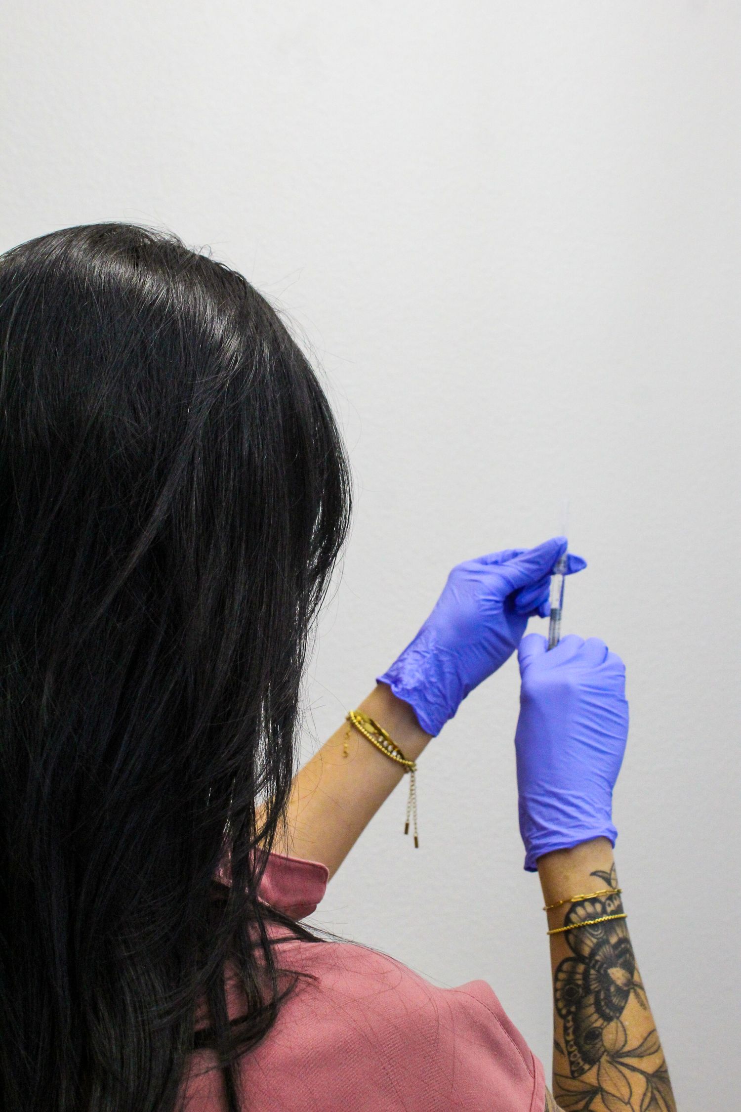 Person wearing blue latex gloves and rose colored shirt performs self injection procedure from behind their shoulder.