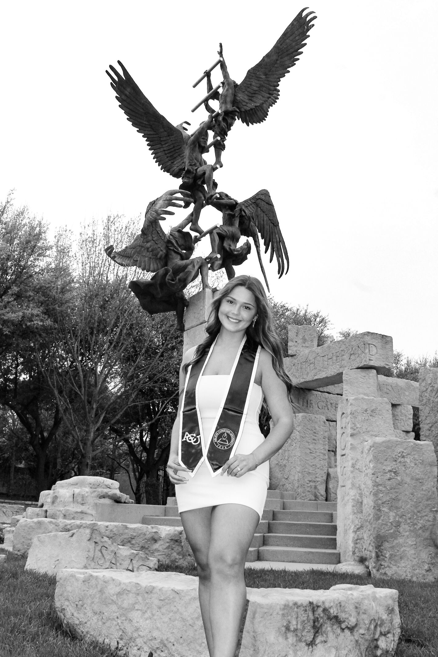 Graduate poses in white dress next to dramatic eagle sculpture on stone steps.
