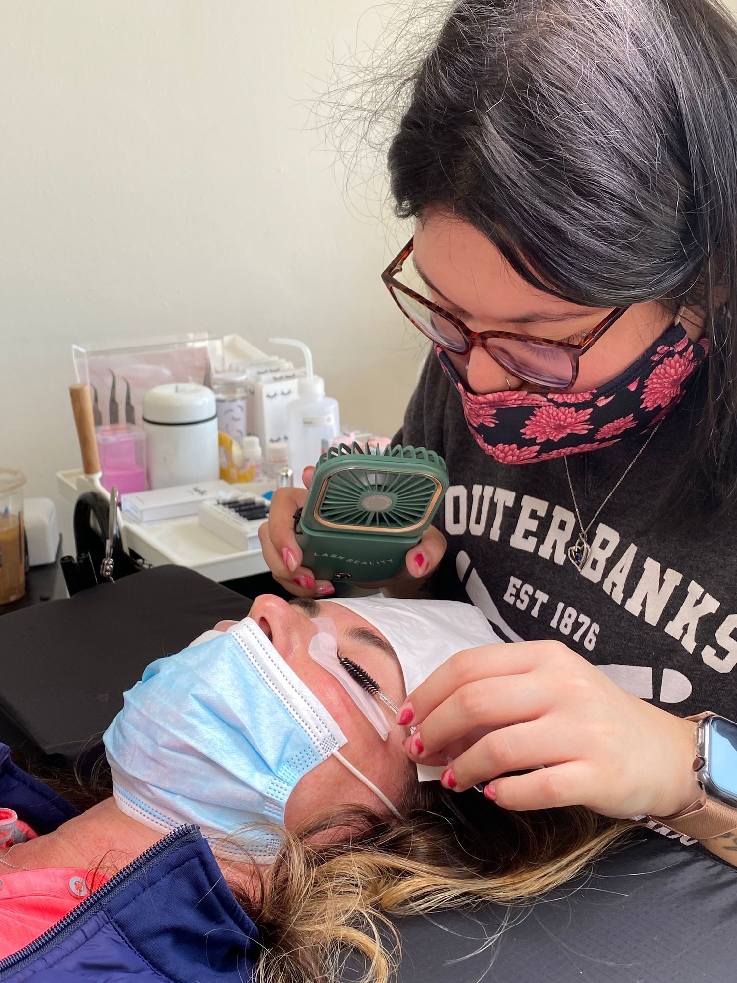 Professional lash technician in glasses and black sweatshirt carefully applies eyelash extensions to client wearing protective mask.