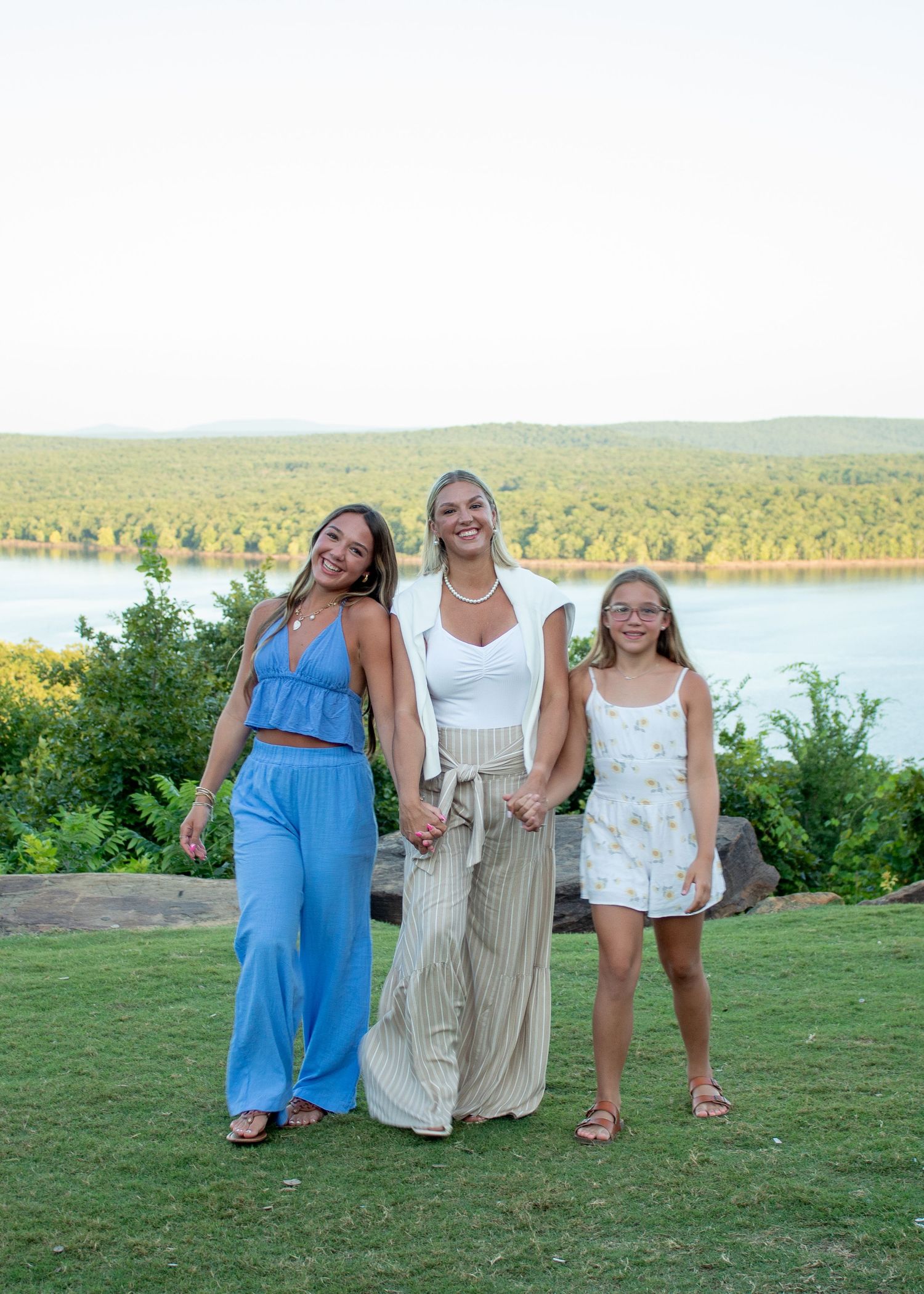 Three people standing together and smiling on grassy hill overlooking scenic lake and forest landscape during sunset.