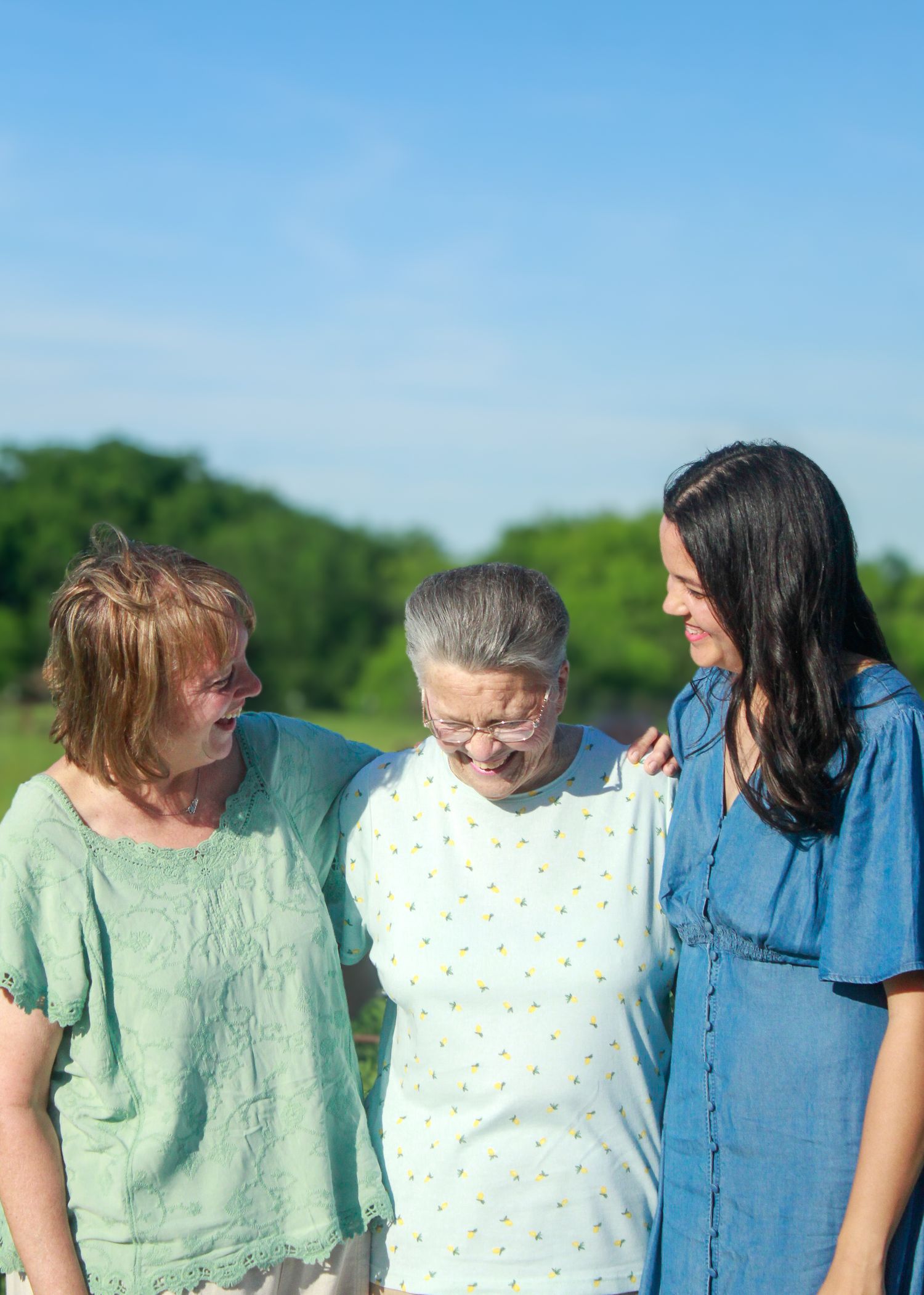 Three women in casual dresses share a joyful moment outdoors on a sunny day.