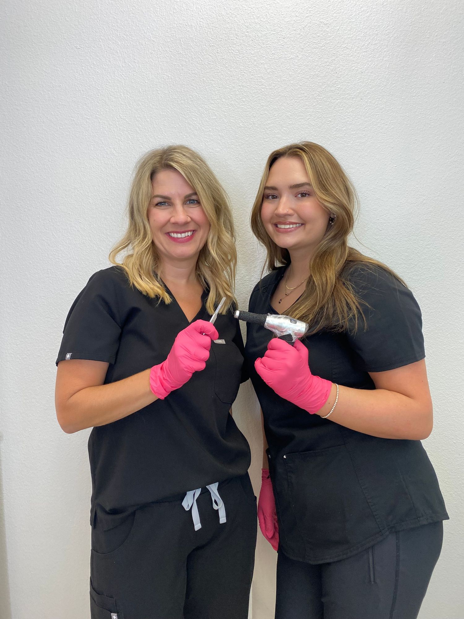 Two healthcare workers in black scrubs and pink gloves smile while posing together against a white wall.