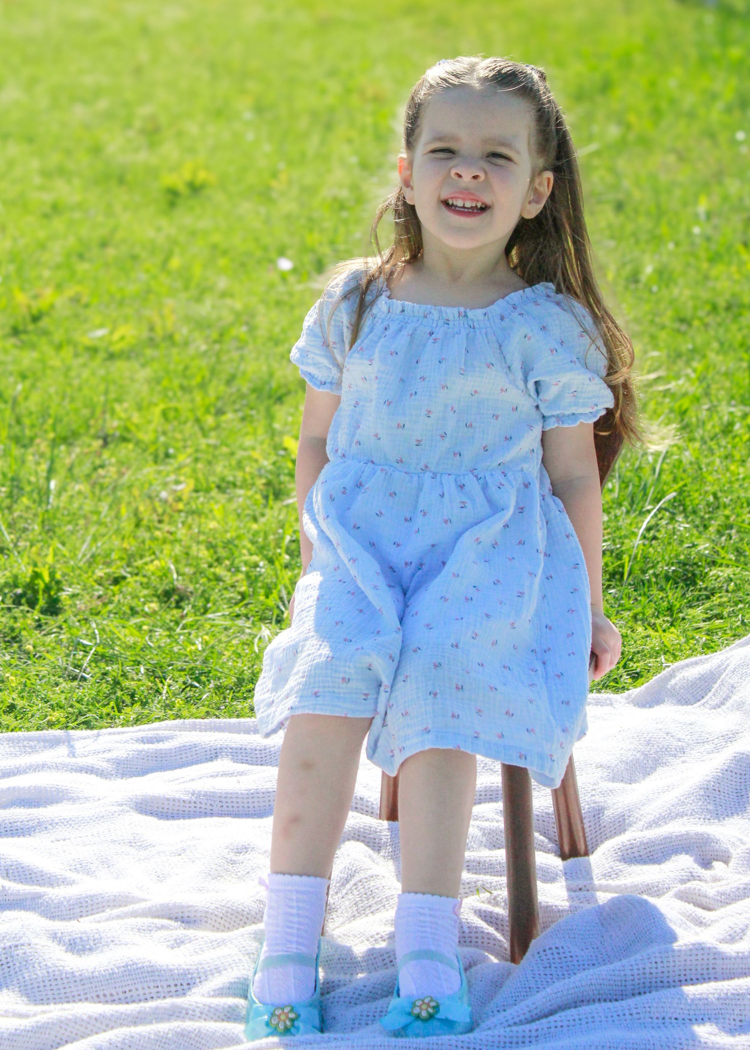 Young child in a light blue dress sits smiling on a picnic blanket in a sunny garden.