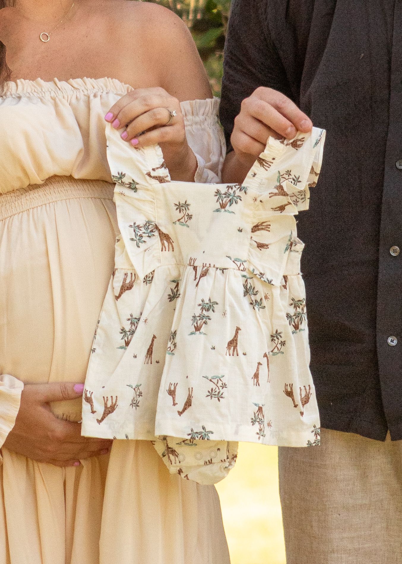Expectant couple holding up a delicate white baby dress with floral and giraffe print against a sunlit outdoor backdrop.