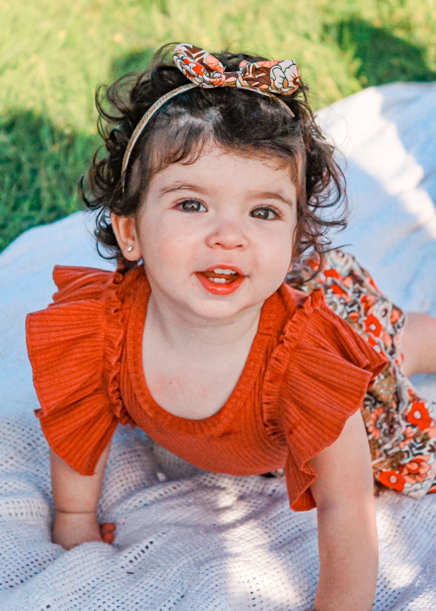Child wearing orange ruffled dress laying on white blanket outdoors.