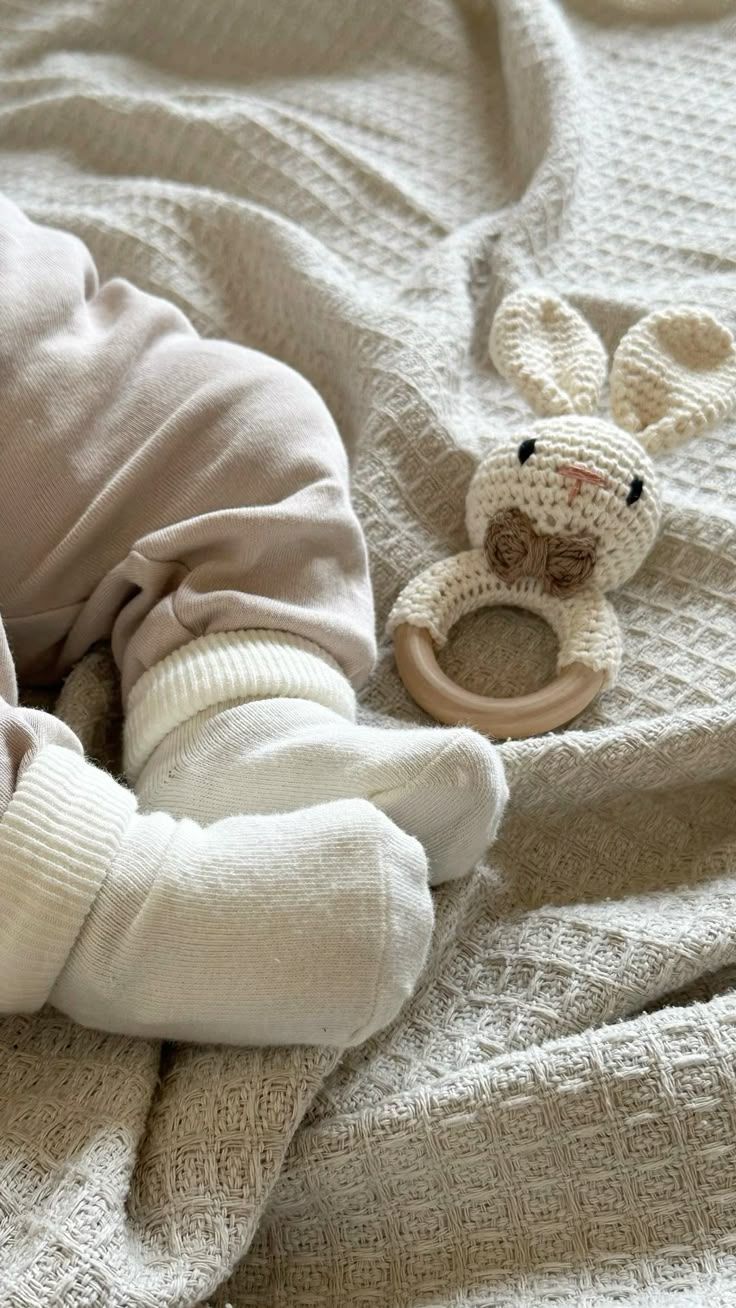 A soft white cable knit blanket with baby feet in socks and a crocheted bunny teething ring resting on top.