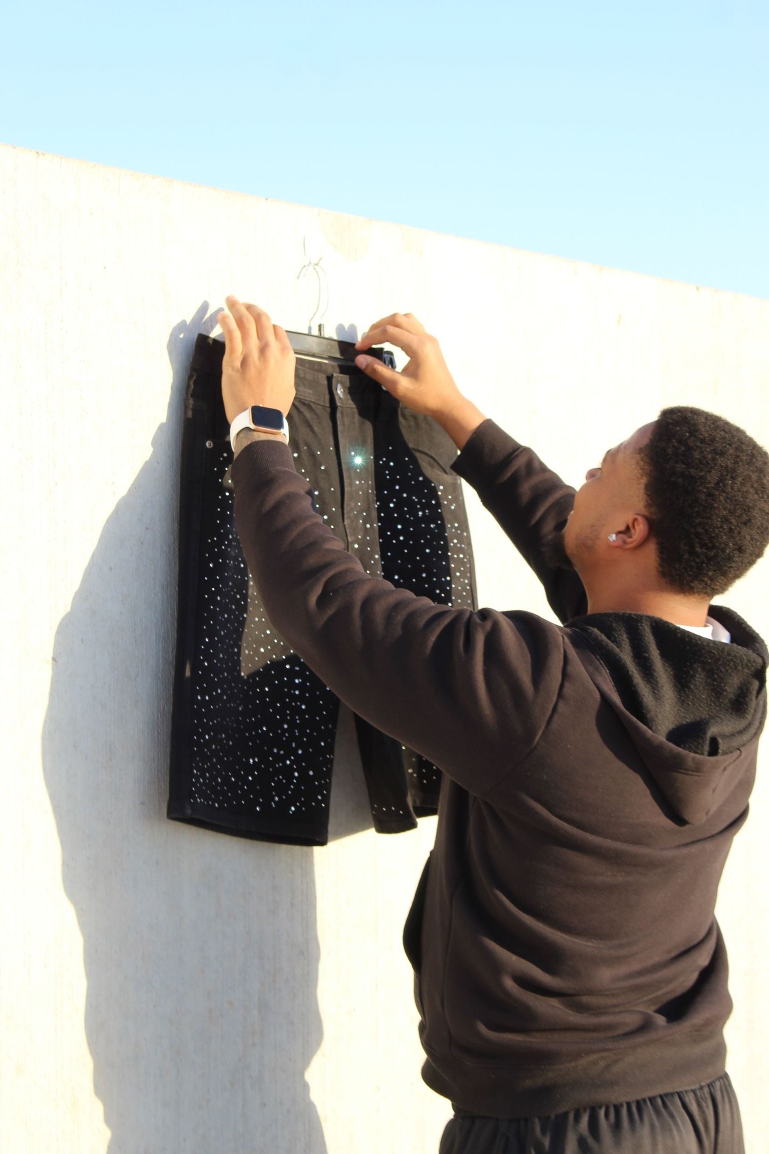 Someone in a brown hoodie hangs up a sparkly black dress or garment against a light-colored wall outdoors.