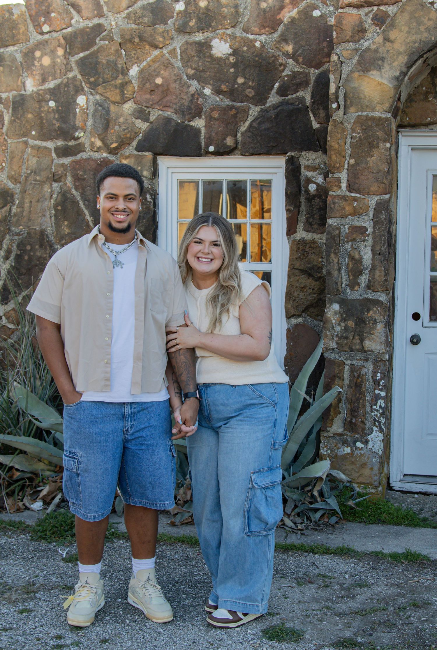 Couple posing together against stone wall wearing casual summer outfits.