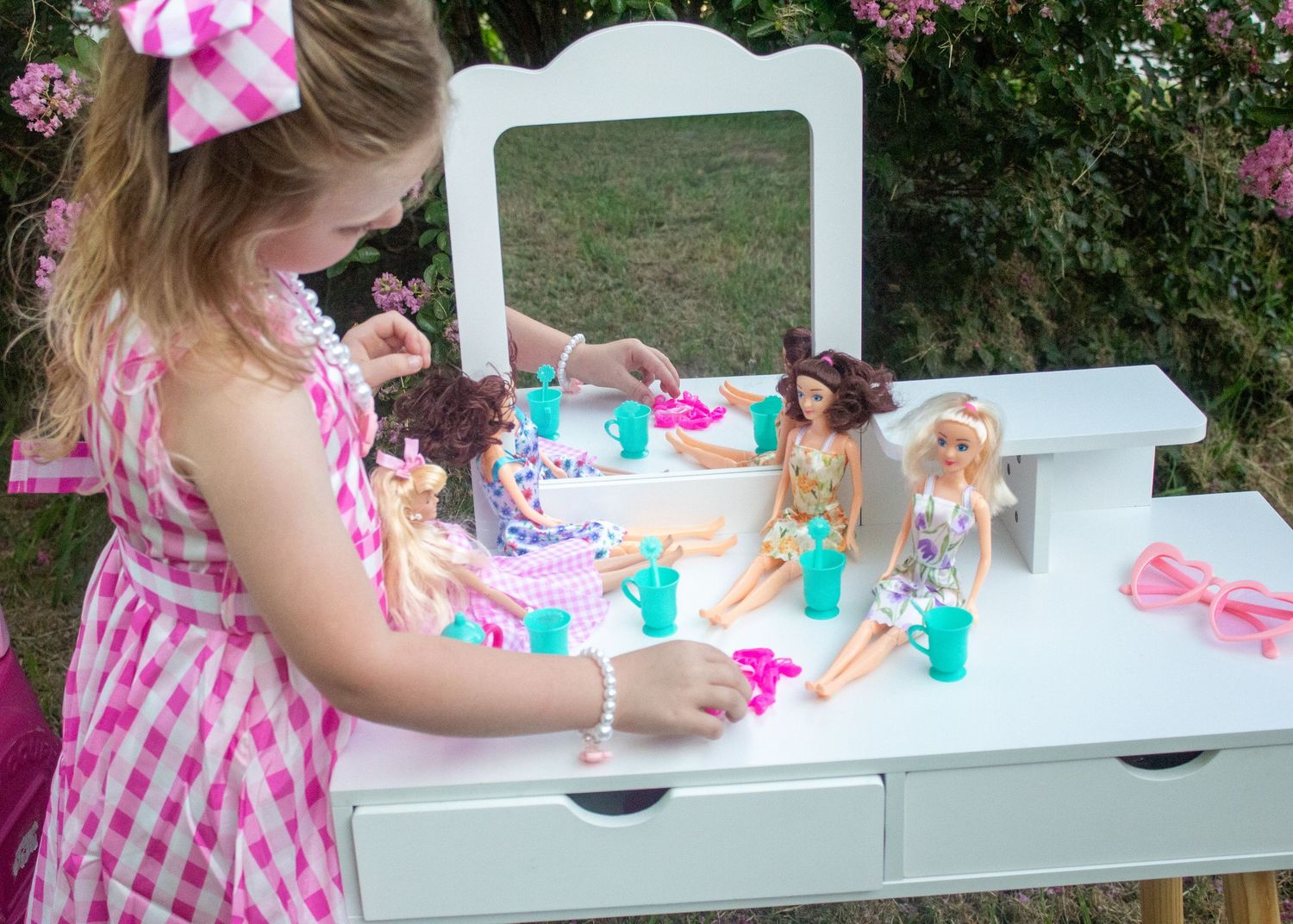 A white wooden tea party vanity table set up outdoors with dolls and tea party accessories in pink and turquoise.