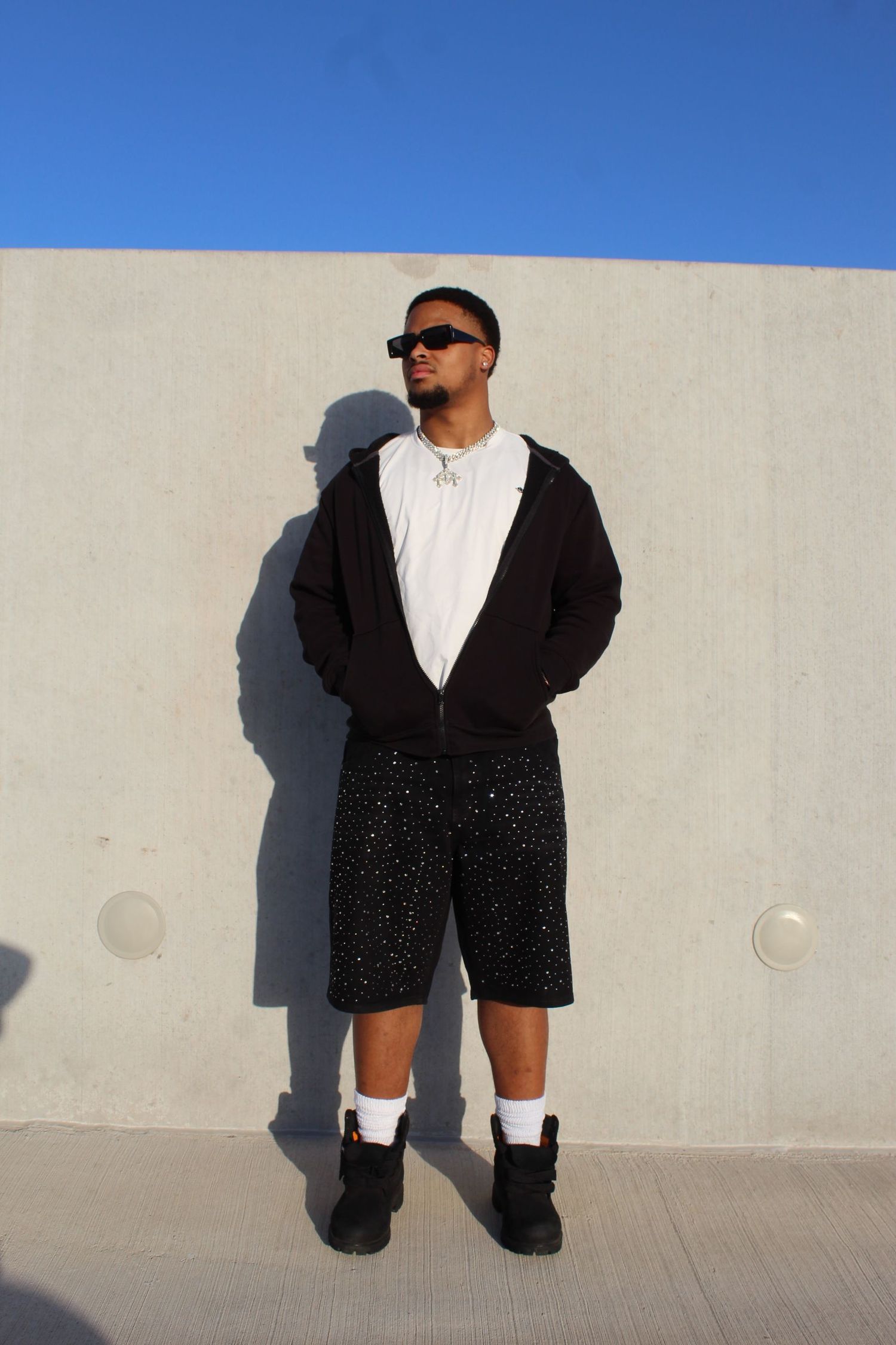 Person in sunglasses wearing black hoodie, white shirt, sparkly shorts and boots poses against concrete wall in sunlight.