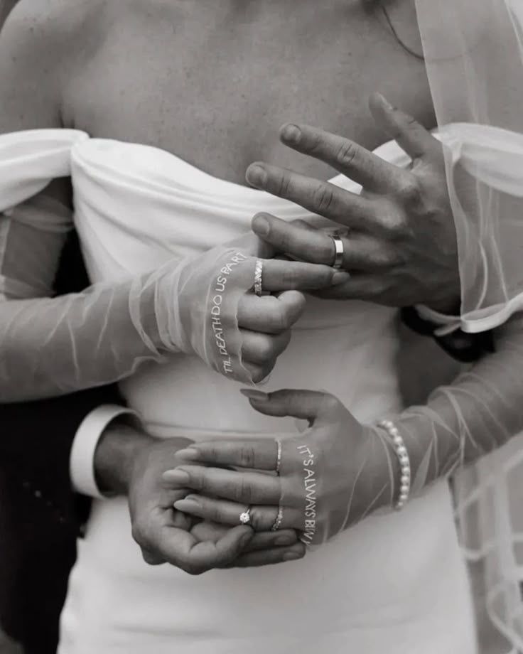 A black and white photograph showing hands with wedding rings embracing through delicate sheer fabric and lace.