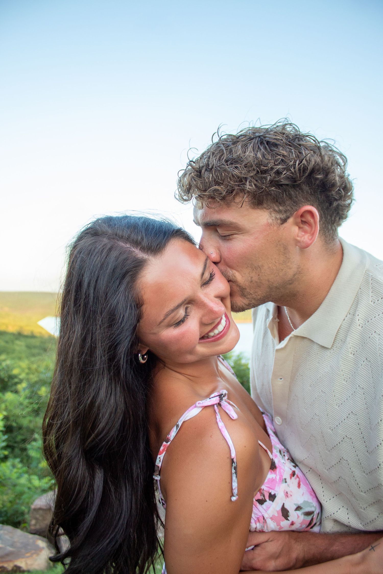 A couple embraces in a romantic photo series set against an outdoor backdrop with blue sky and greenery.