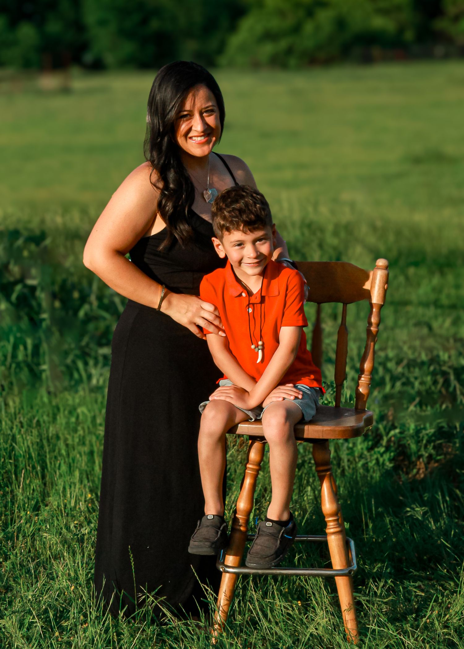 A sweet outdoor portrait with someone in black dress and a child sitting on wooden chair in grassy field.