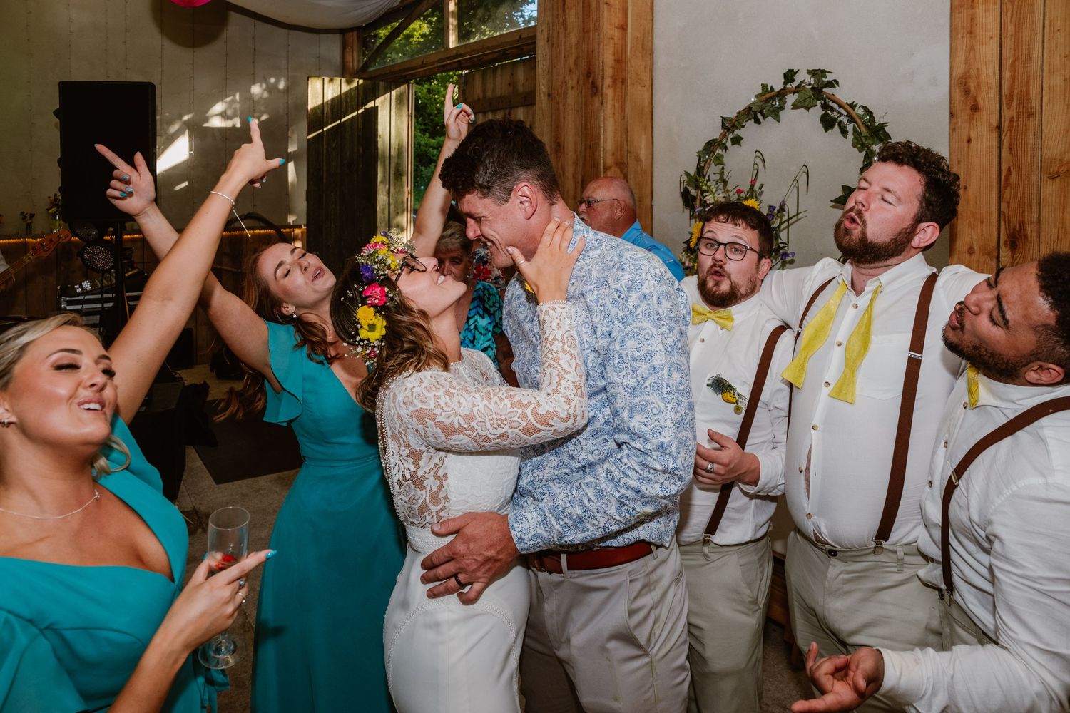 Bride and groom share romantic dance surrounded by cheering wedding guests in teal dresses and suspenders.