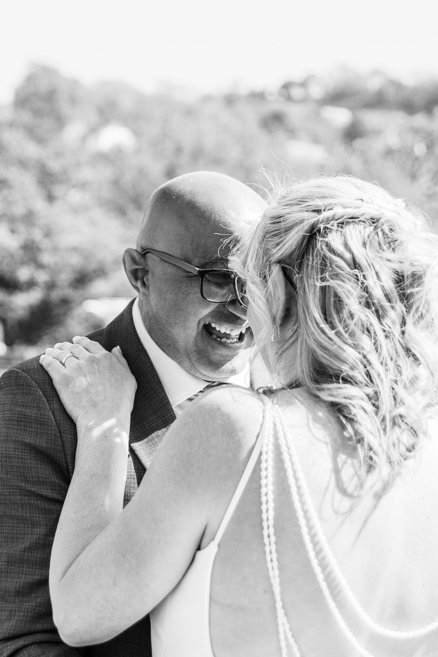 Black and white photo of a bald groom laughing joyfully while embracing a blonde bride in a pearl-back wedding dress.