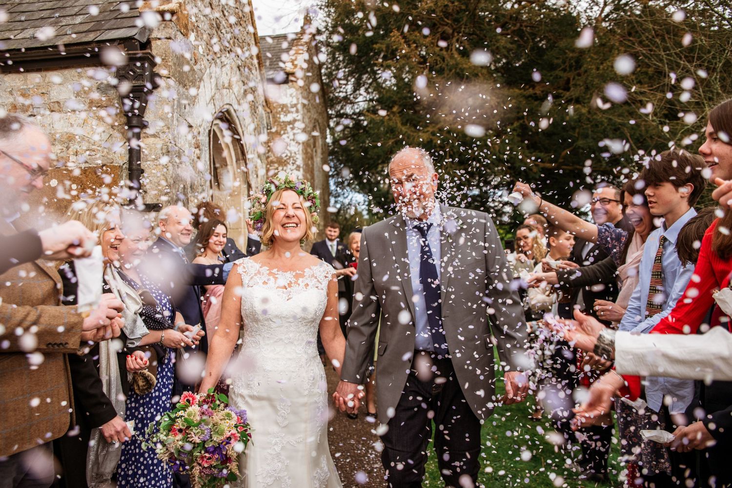 Bride and groom walk through confetti shower outside historic church surrounded by cheering wedding guests.
