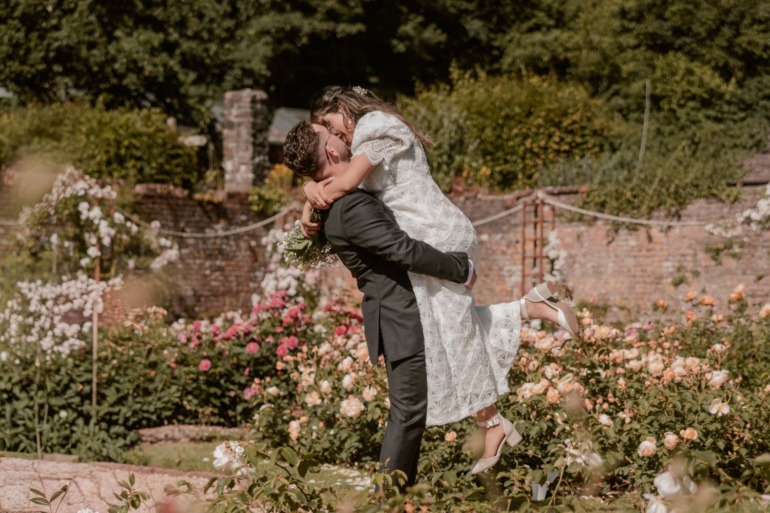 Groom lifting bride in a romantic garden filled with pink and white roses during a wedding celebration.