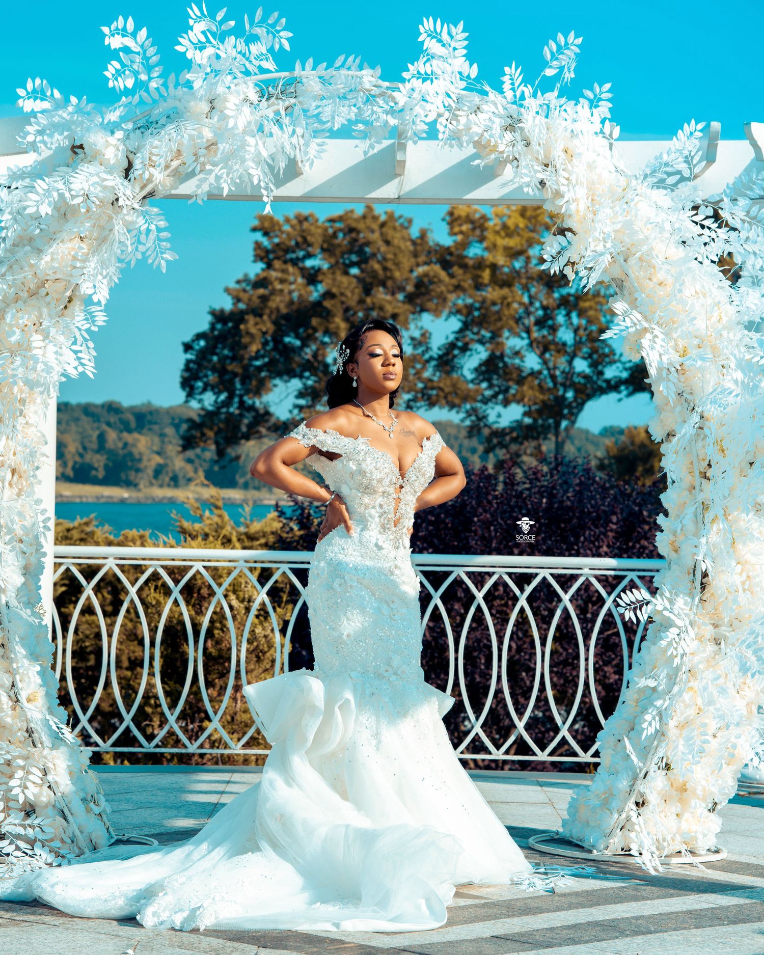 Bride in mermaid wedding dress poses under white floral arch against scenic waterfront backdrop.