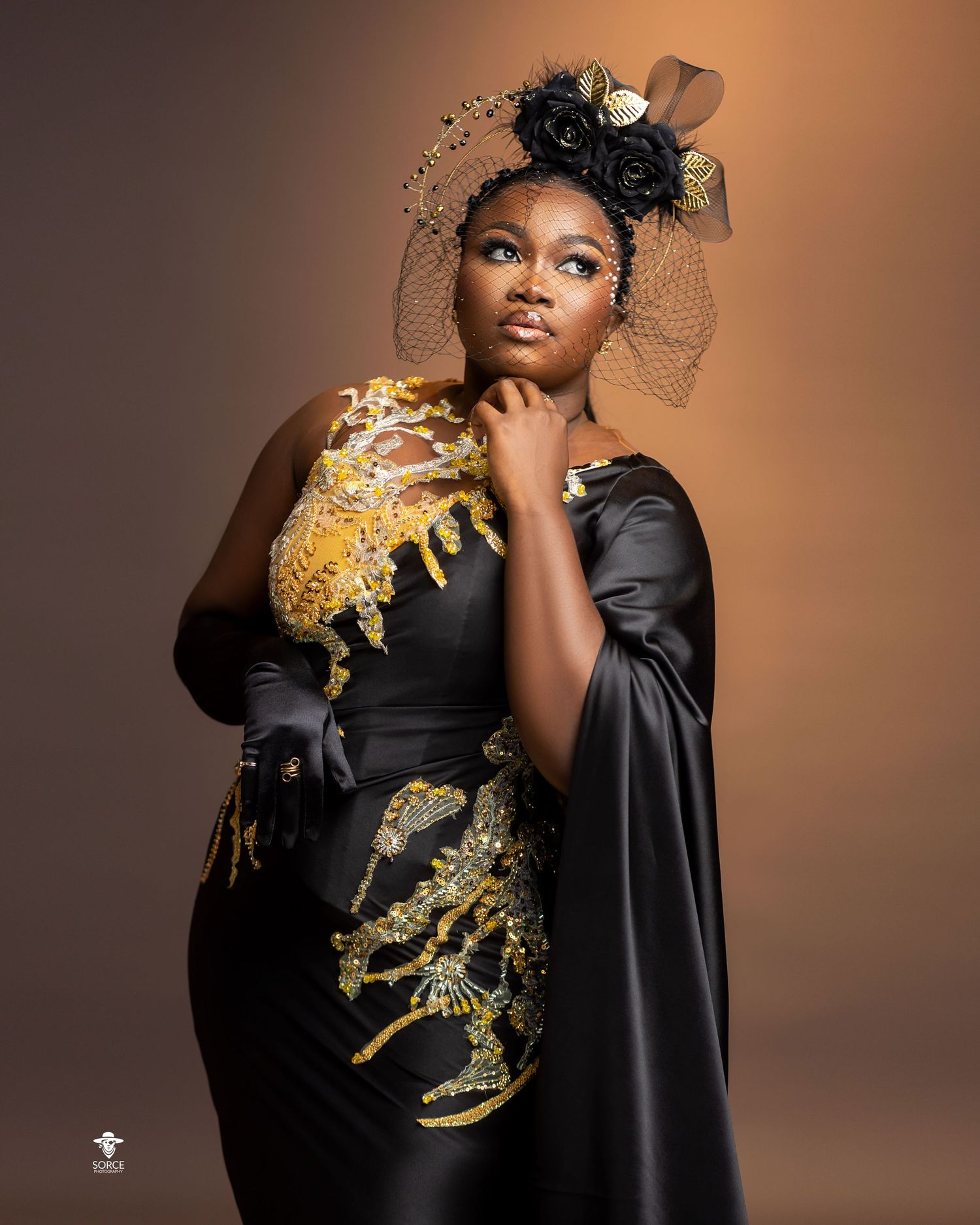 Fashion portrait in black satin dress with dramatic gold embellishments against warm brown background.