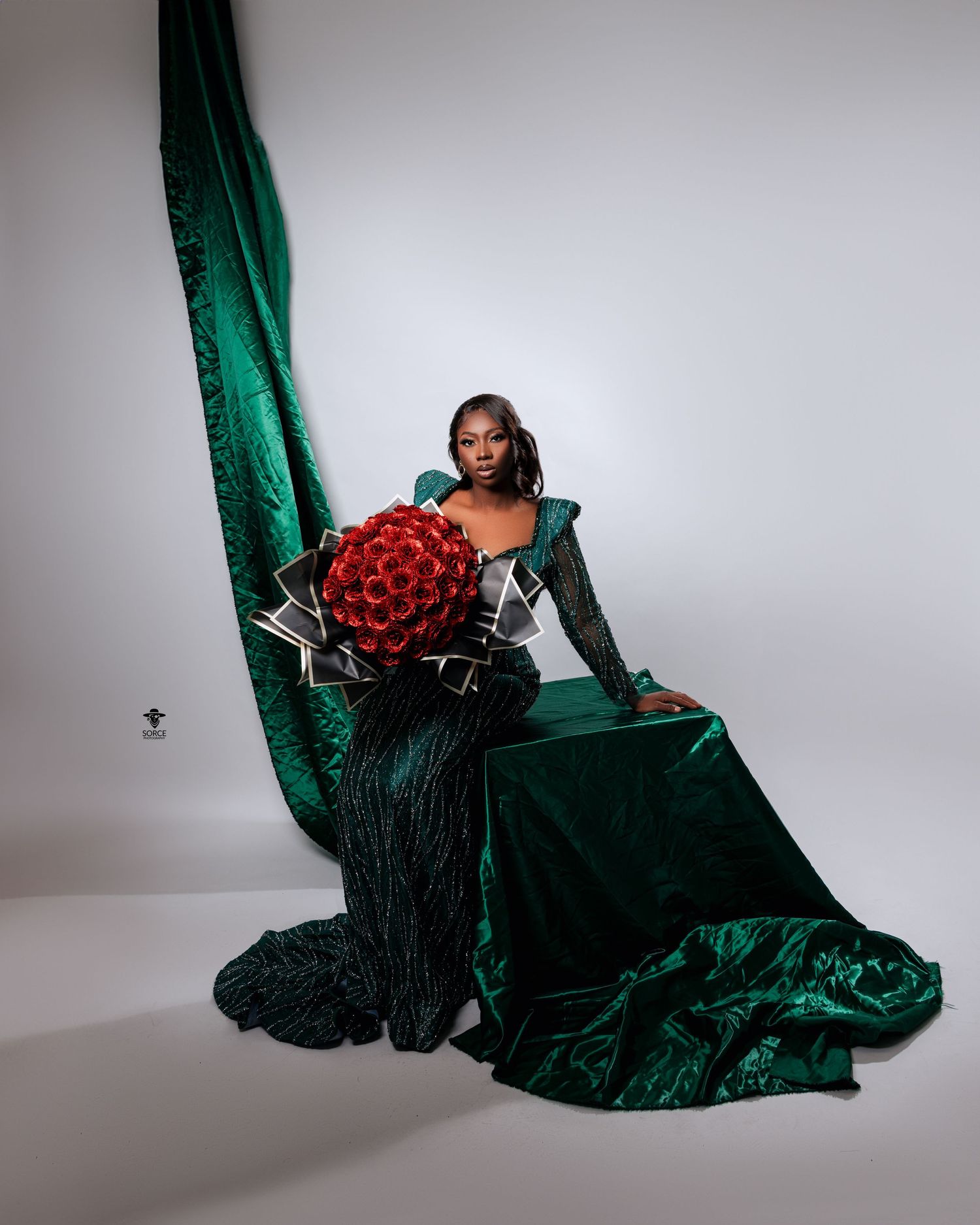 Elegant fashion portrait against white background featuring green velvet gown and vibrant red rose bouquet.