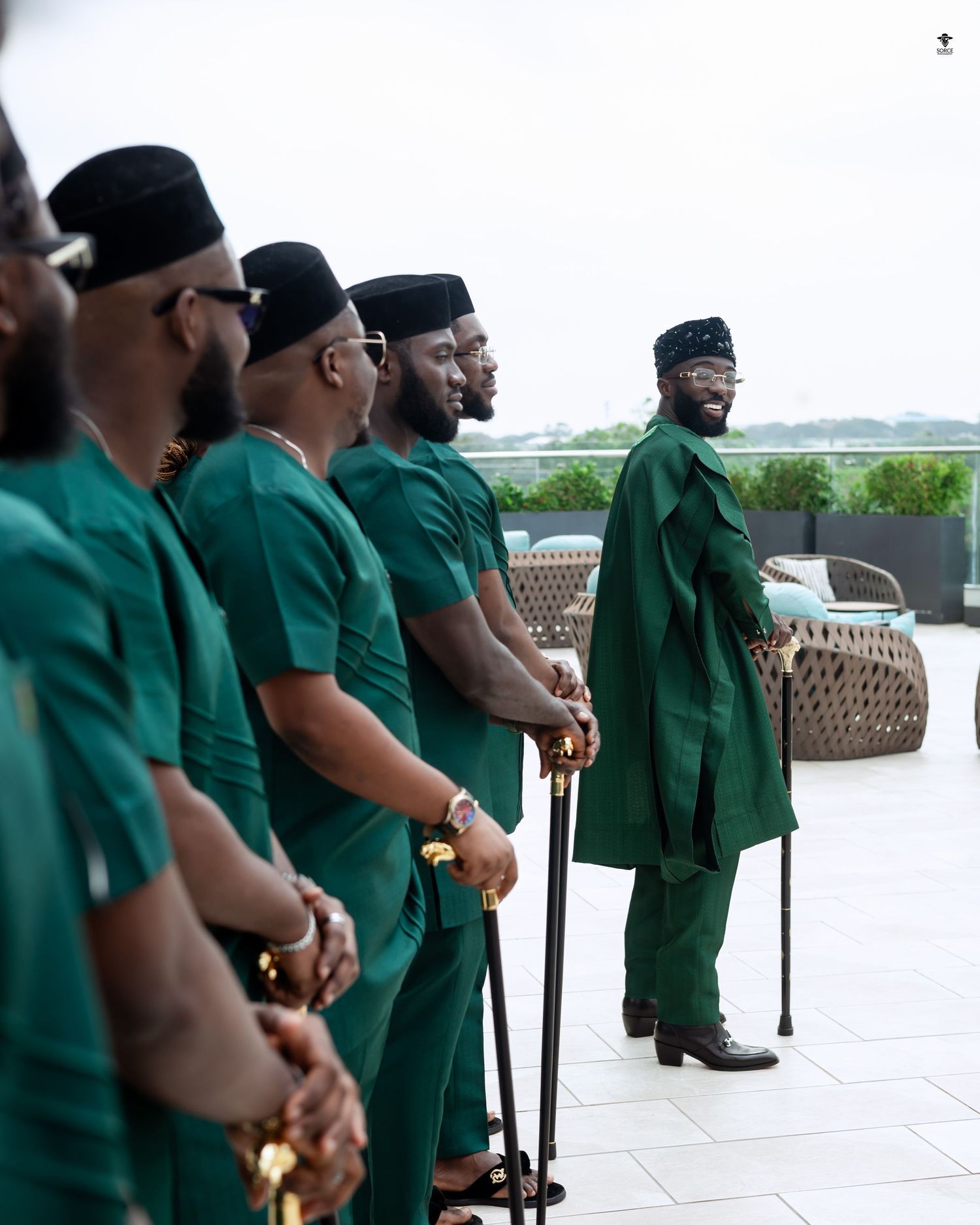 Wedding party wearing matching green traditional outfits stand in line on an outdoor patio with canes.