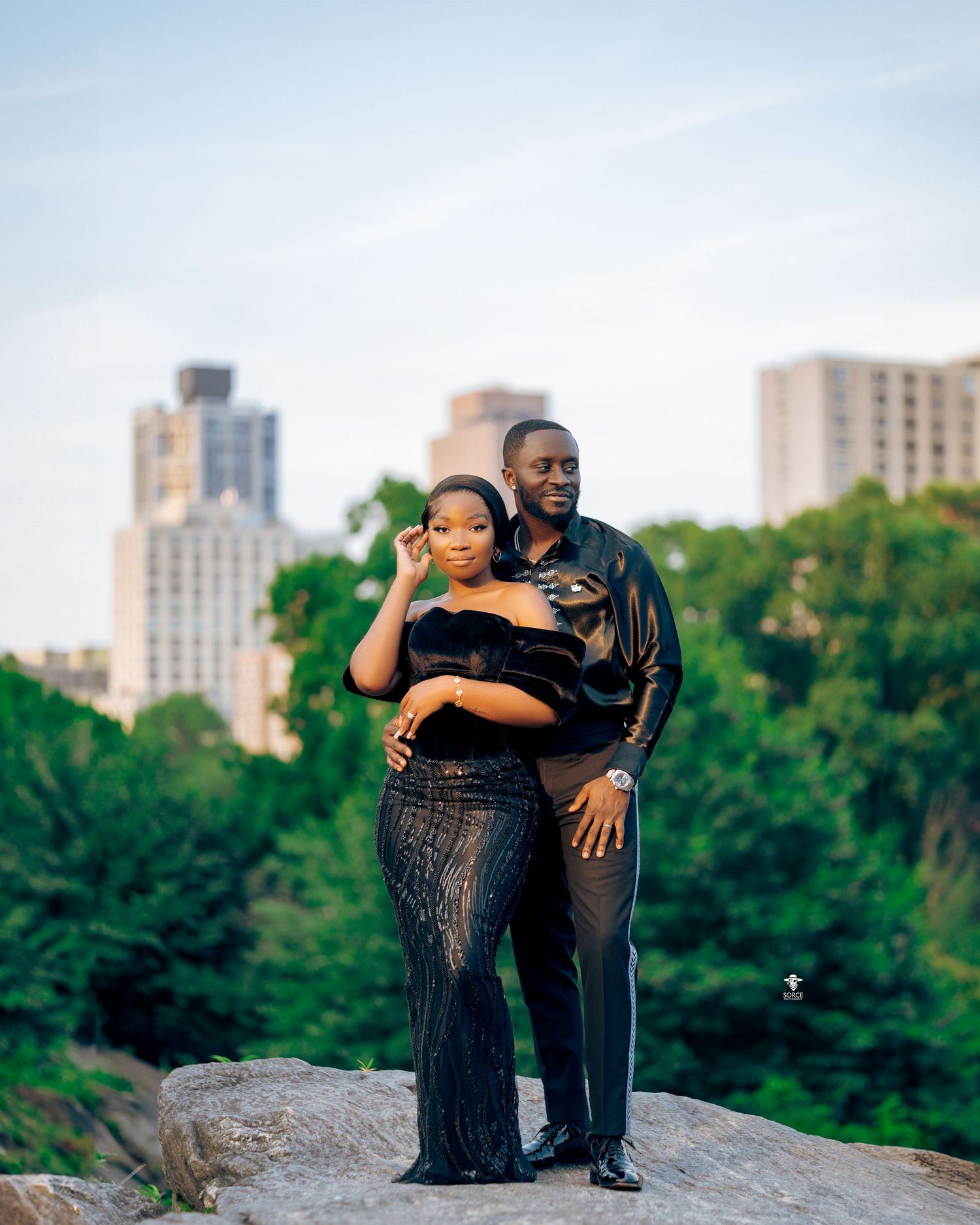 A romantic couple embraces while standing on a rock outcrop with city buildings and green trees in the background.