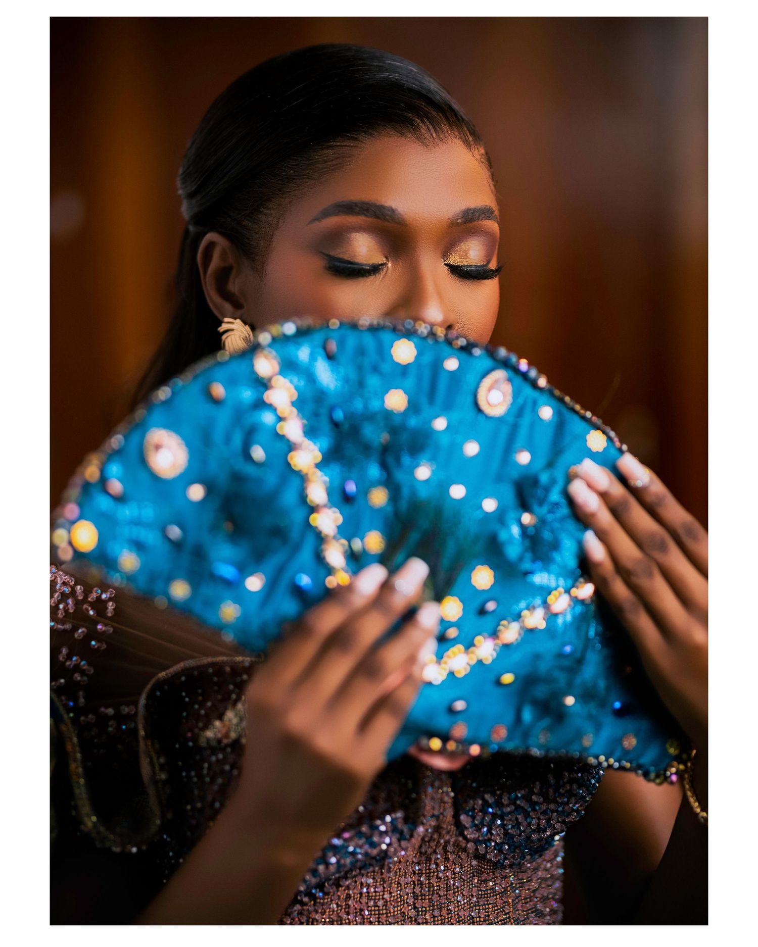 A stunning portrait with a decorative blue fan adorned with pearls and sparkles against a dark background.