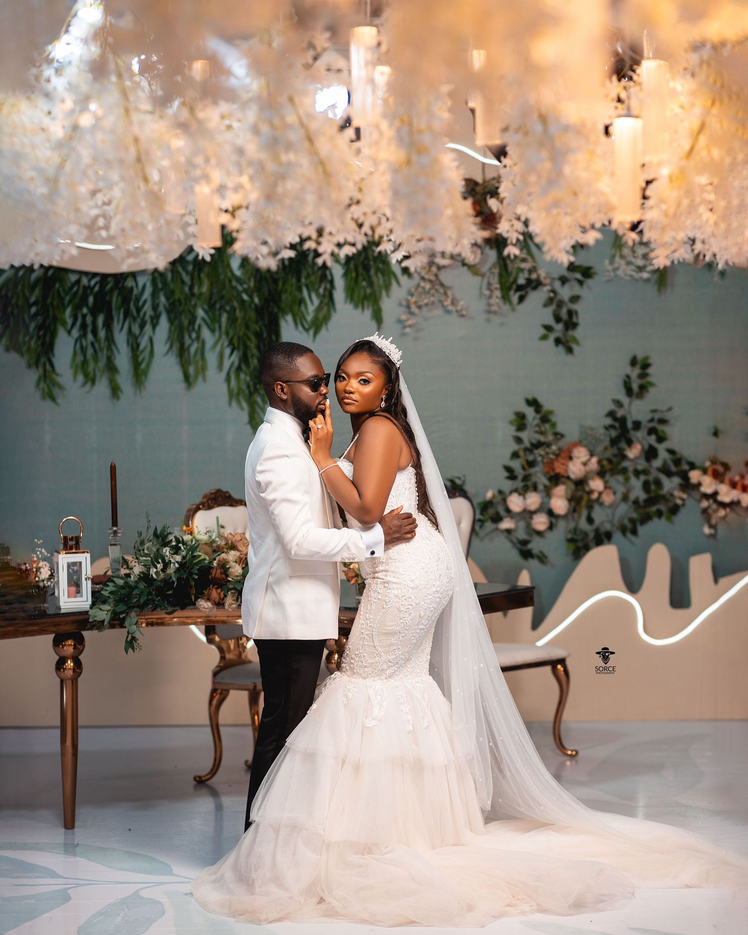 A wedding couple embraces in a beautifully decorated indoor venue with hanging lights and greenery against a light wall.