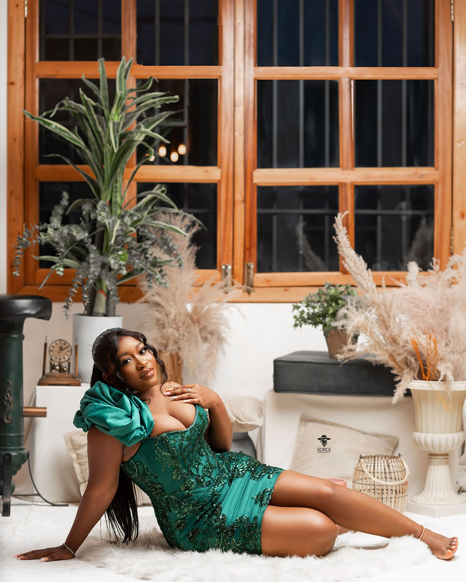 Woman in emerald green sequin dress poses elegantly against white modern interior with plants and windows.