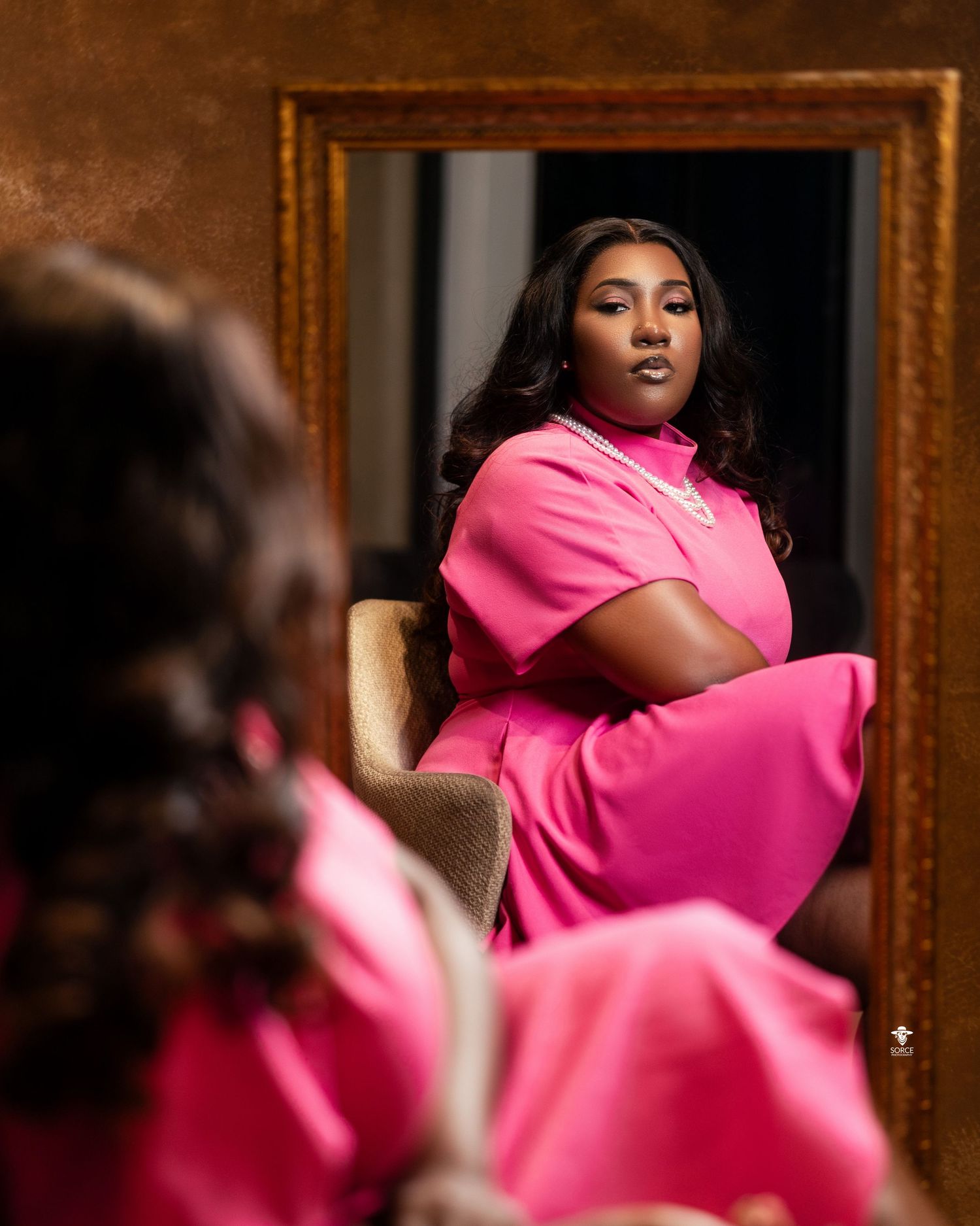 A reflection in a gold-framed mirror shows someone wearing a vibrant hot pink outfit while seated in an elegant chair.