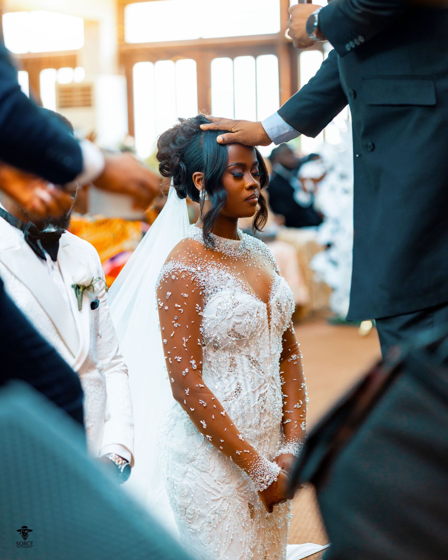 A person in an elegant white lace wedding gown with sheer sleeves stands surrounded by others during a ceremony.