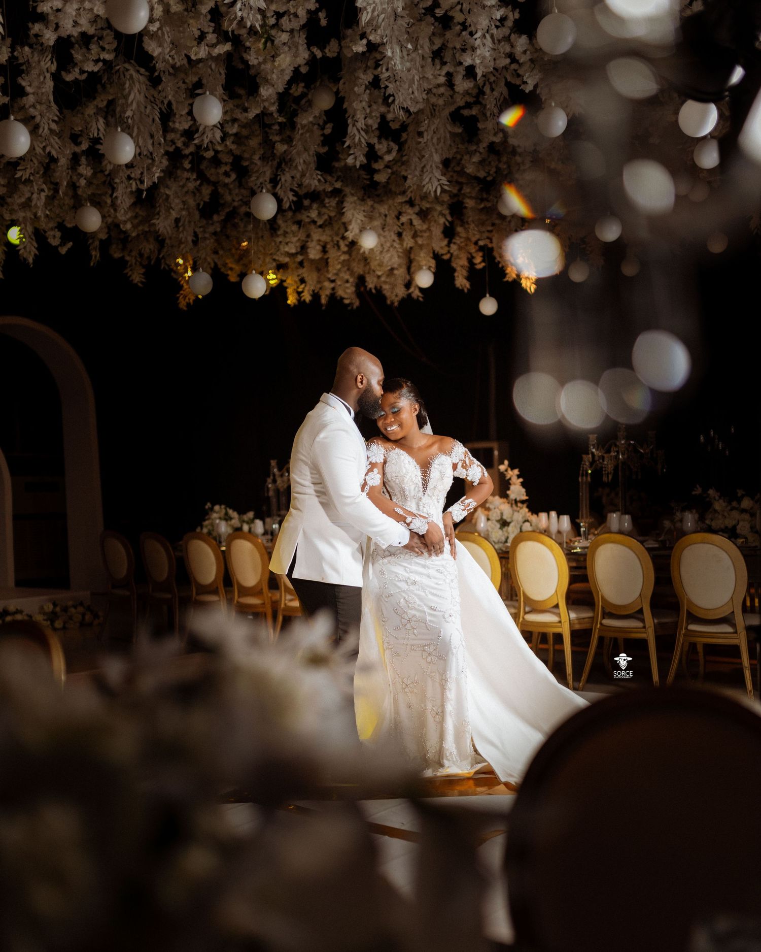 A couple shares an intimate dance moment under twinkling lights at their elegant white wedding reception.