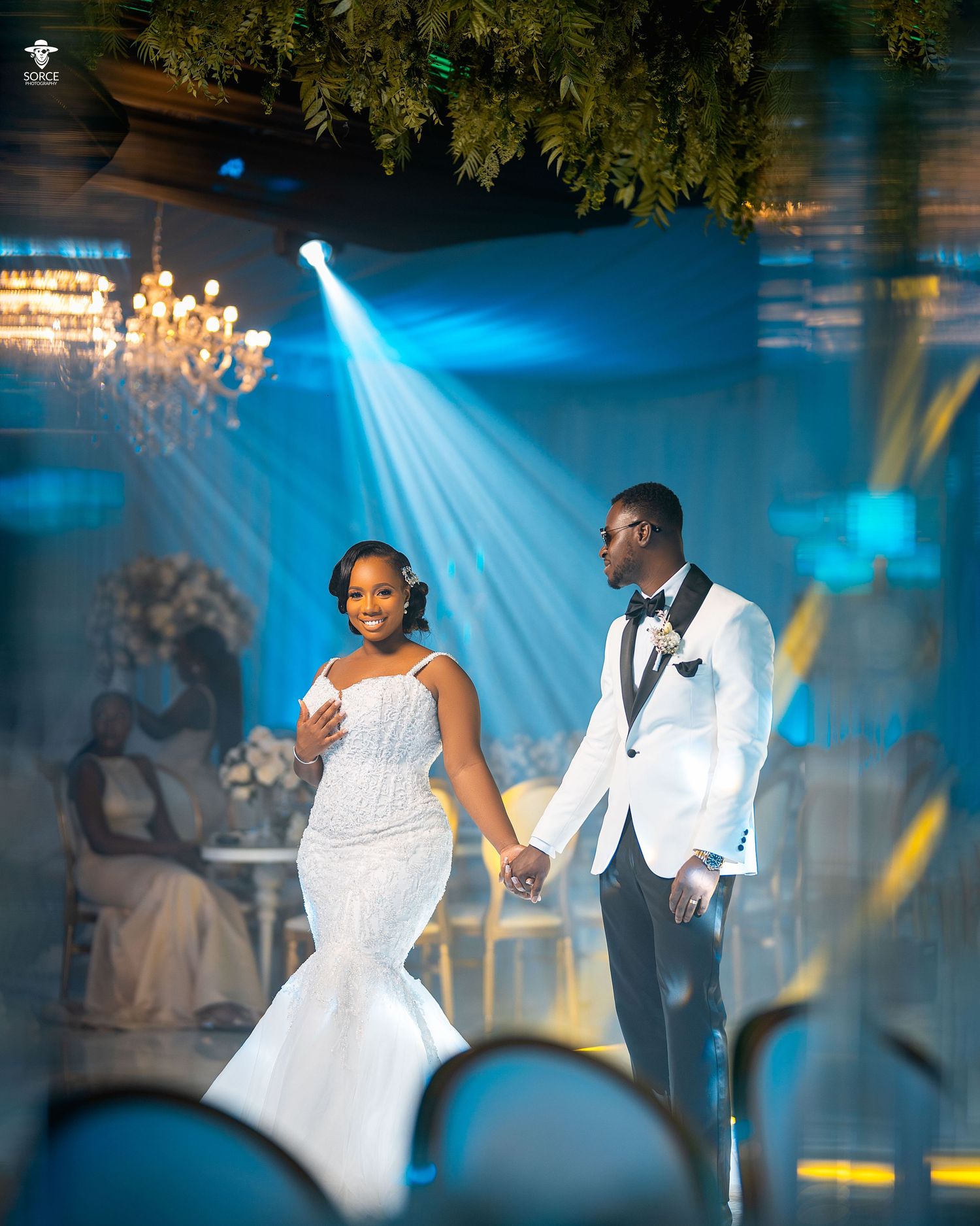 A couple shares their first dance at a wedding reception under dramatic blue lighting and tree branch decorations.