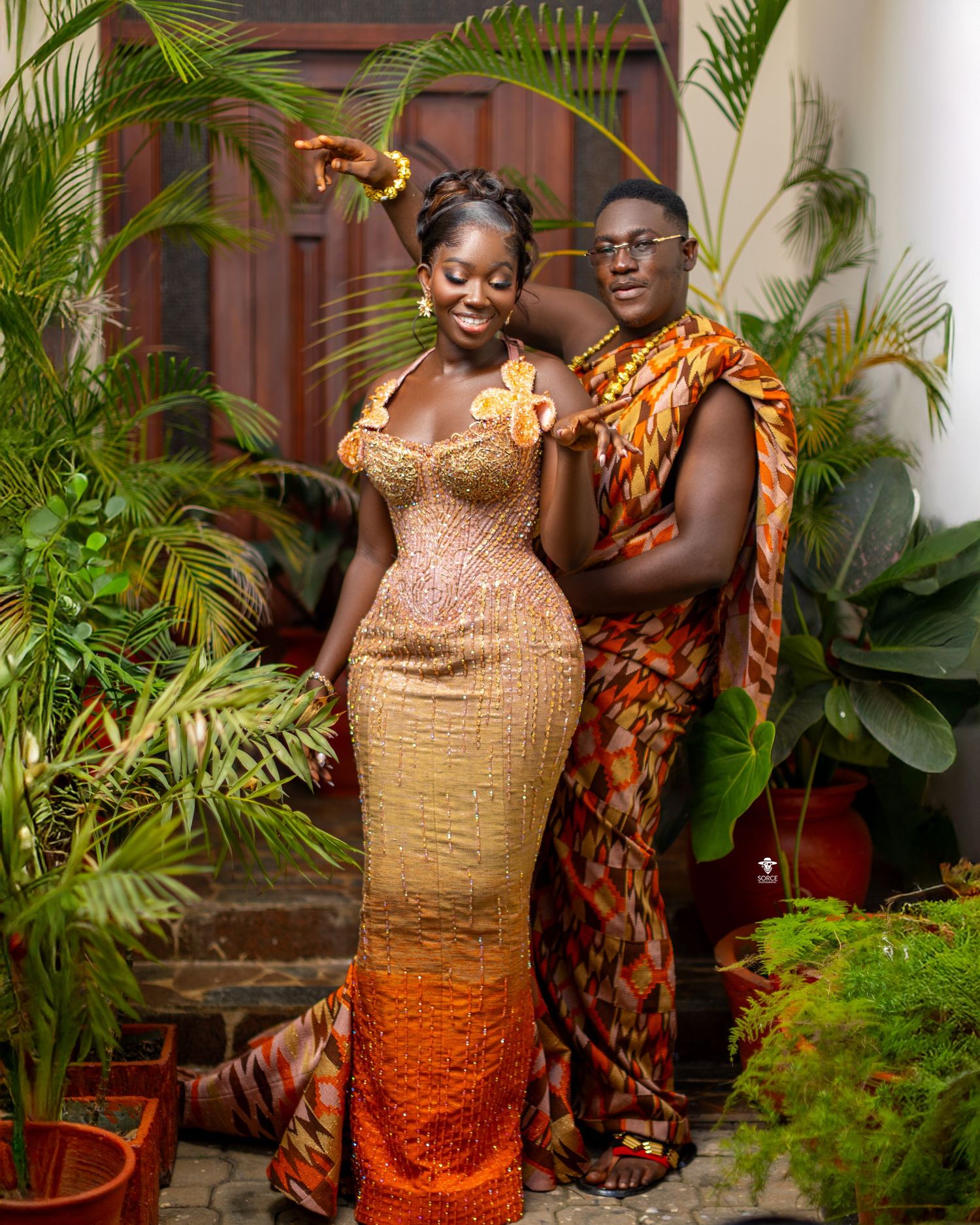 Two people wearing traditional African attire pose together against a backdrop of tropical plants and wooden doors.
