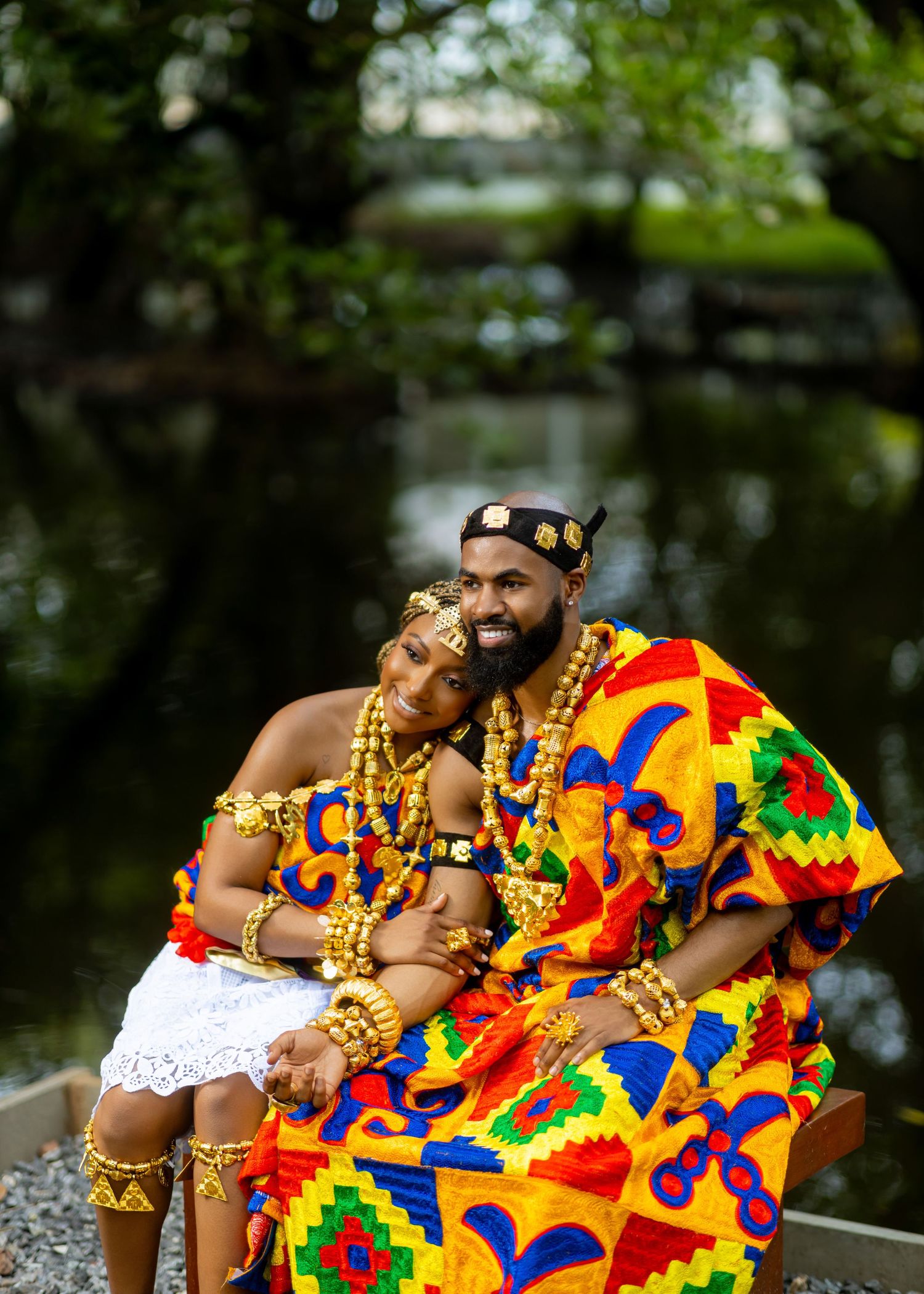 Traditional African couple wearing vibrant Kente cloth with gold beaded jewelry sit together in an outdoor garden setting.