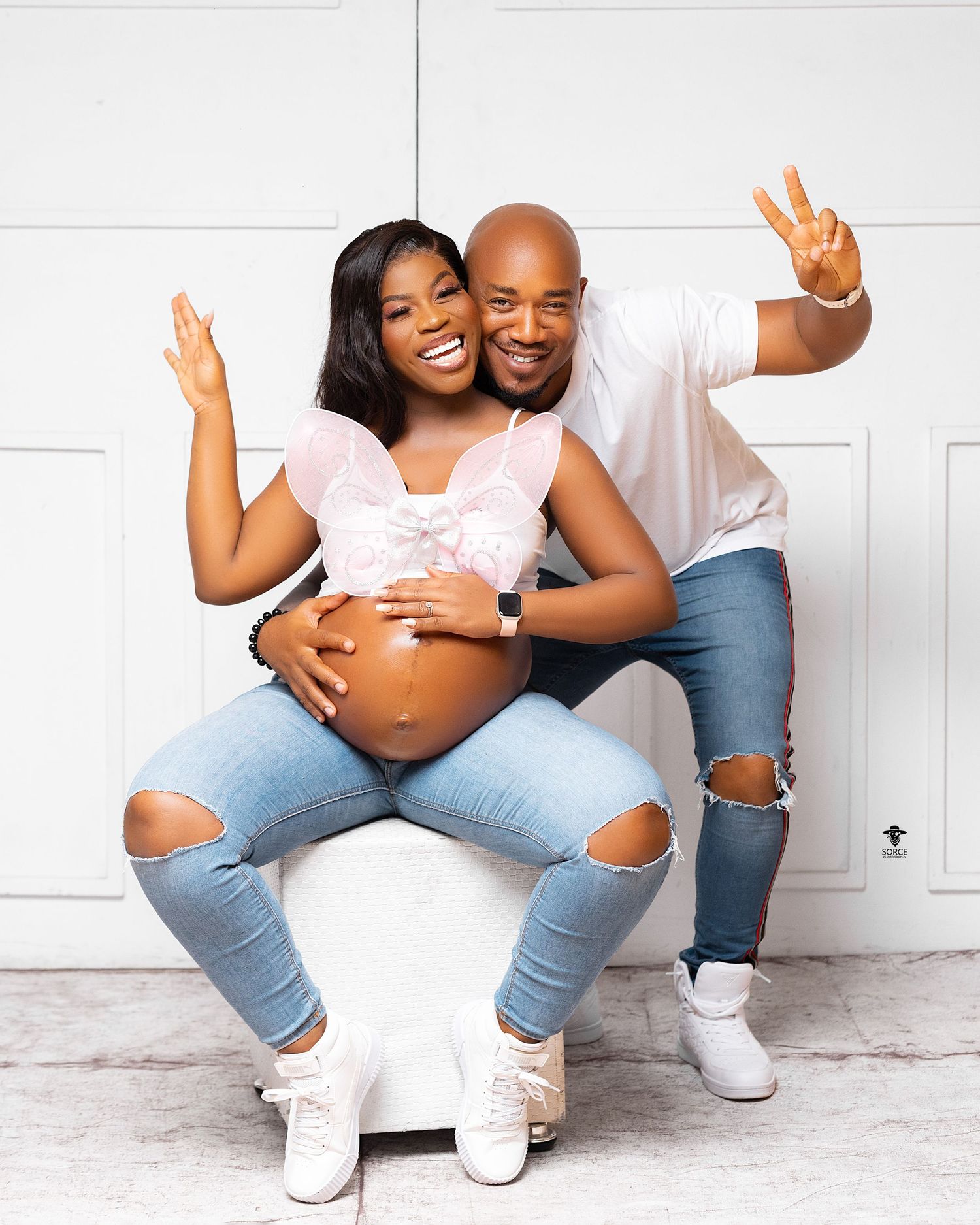 Couple in ripped jeans and white tops poses together against white wall showing excitement and love in maternity photos.