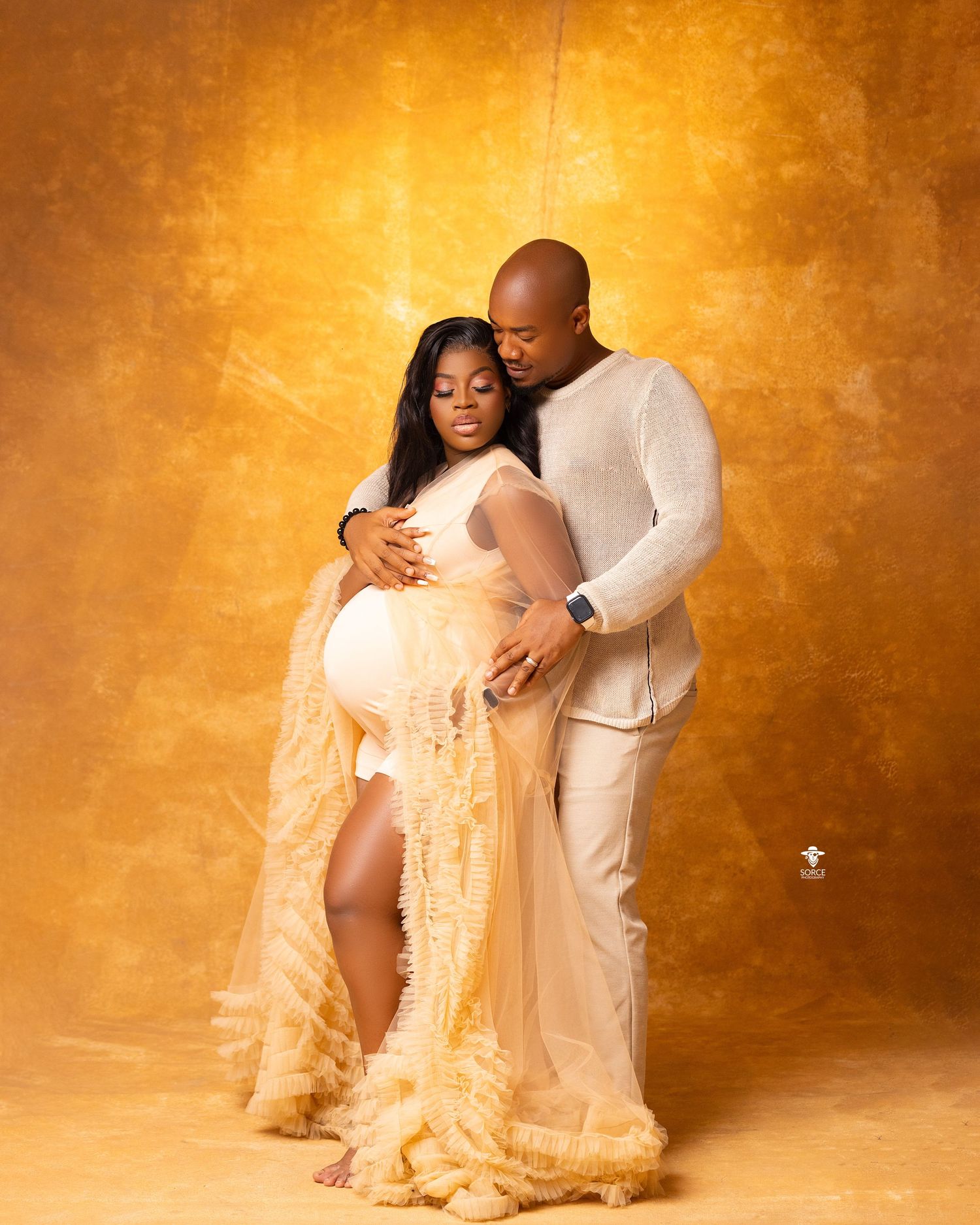 A romantic maternity photoshoot features a couple in white clothing against a warm golden backdrop.