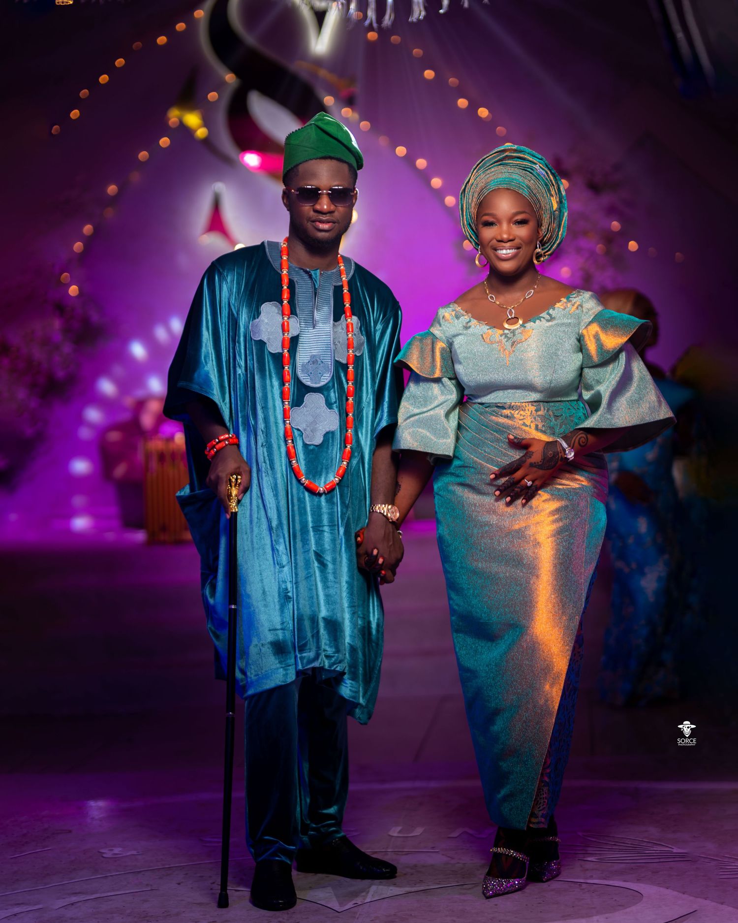 A couple in matching blue and turquoise traditional African attire dance together on a purple-lit stage during a celebration.