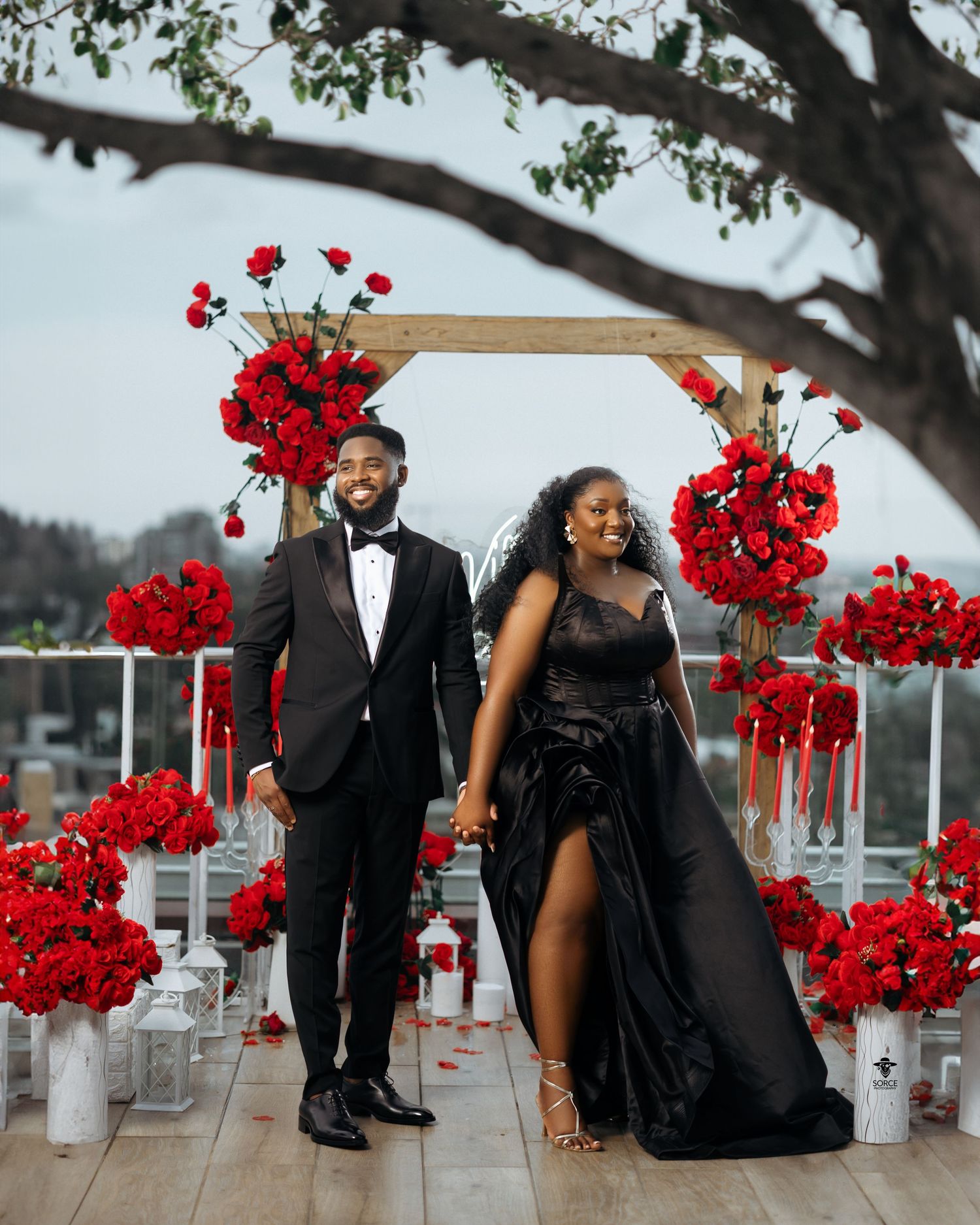 Elegant couple in black formal wear posing before a red rose floral arch at an outdoor wedding ceremony.