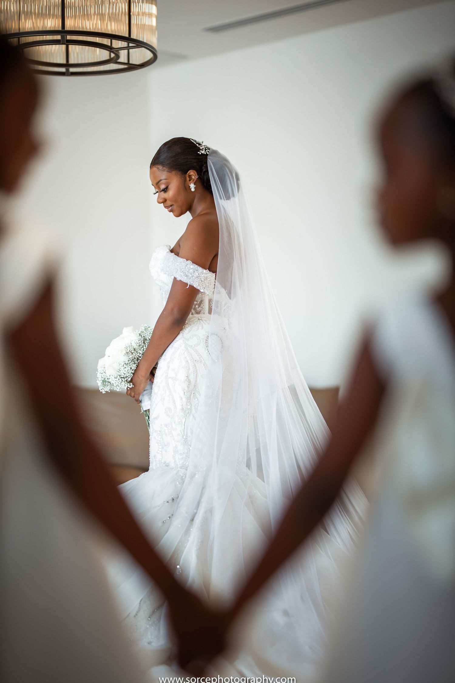 Bride in off-shoulder white wedding gown holding bouquet while being photographed through creative framing.