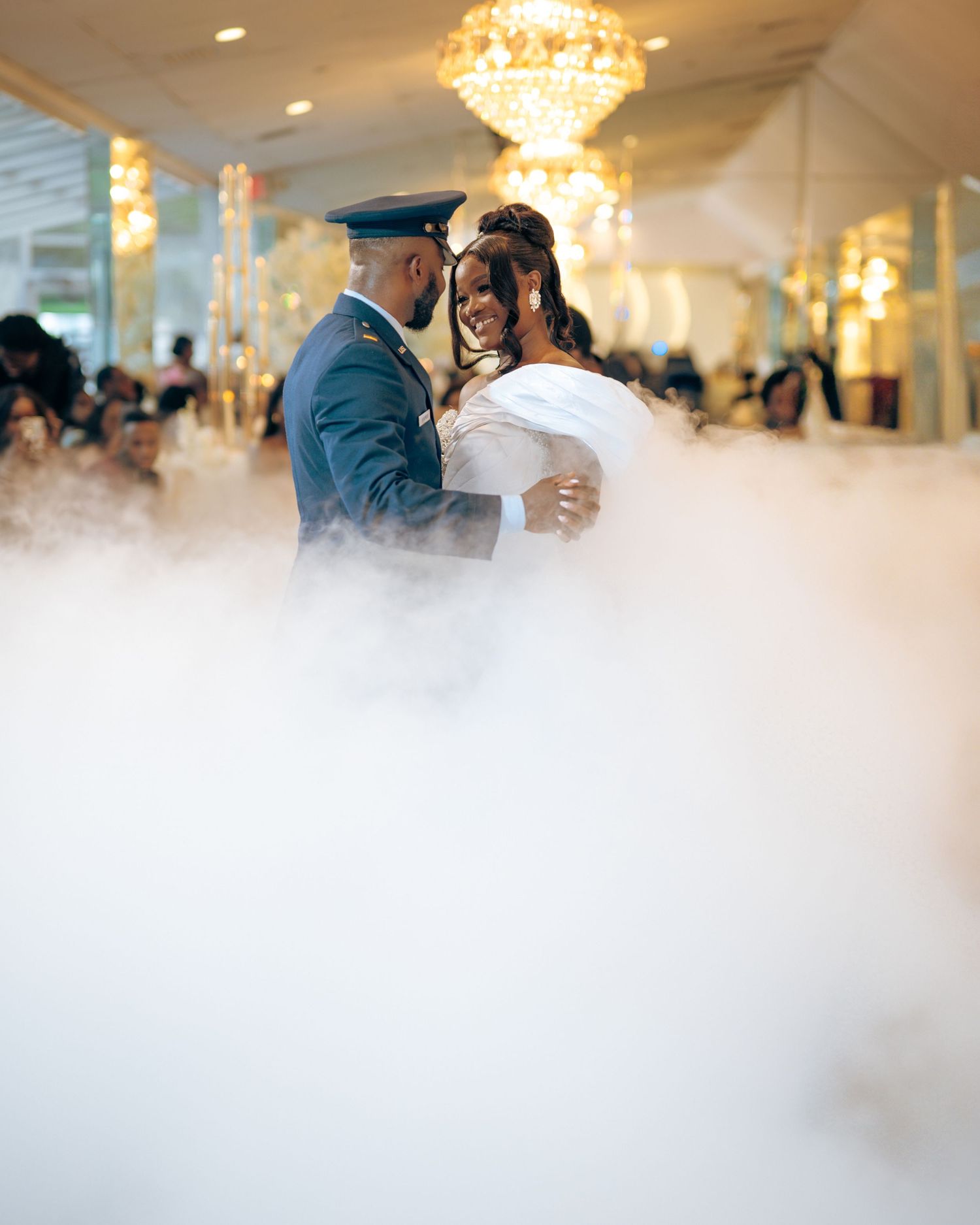 Newlyweds share their first dance surrounded by romantic fog effects and crystal chandeliers at an elegant reception venue.