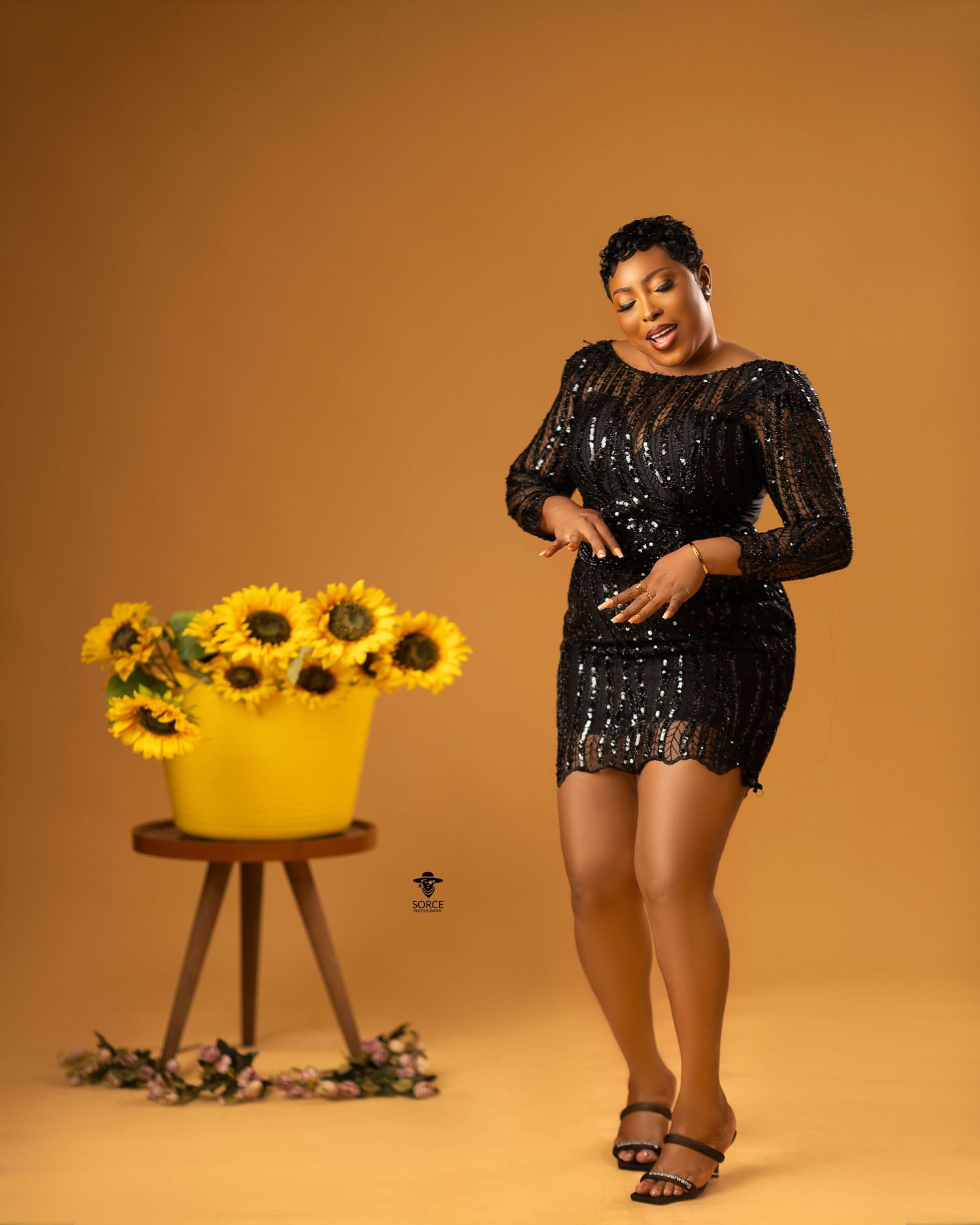 A fashion photo series featuring someone in a sparkling black mini dress posing with sunflowers against an orange backdrop.
