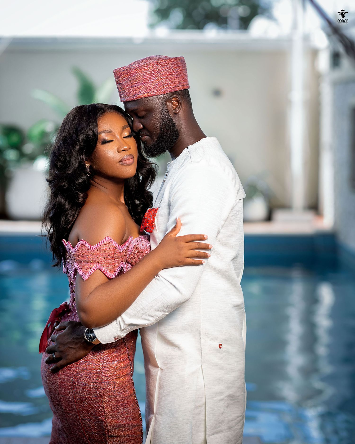 A couple embraces by a pool, with one person wearing a white traditional outfit with red cap and the other in a pink dress.
