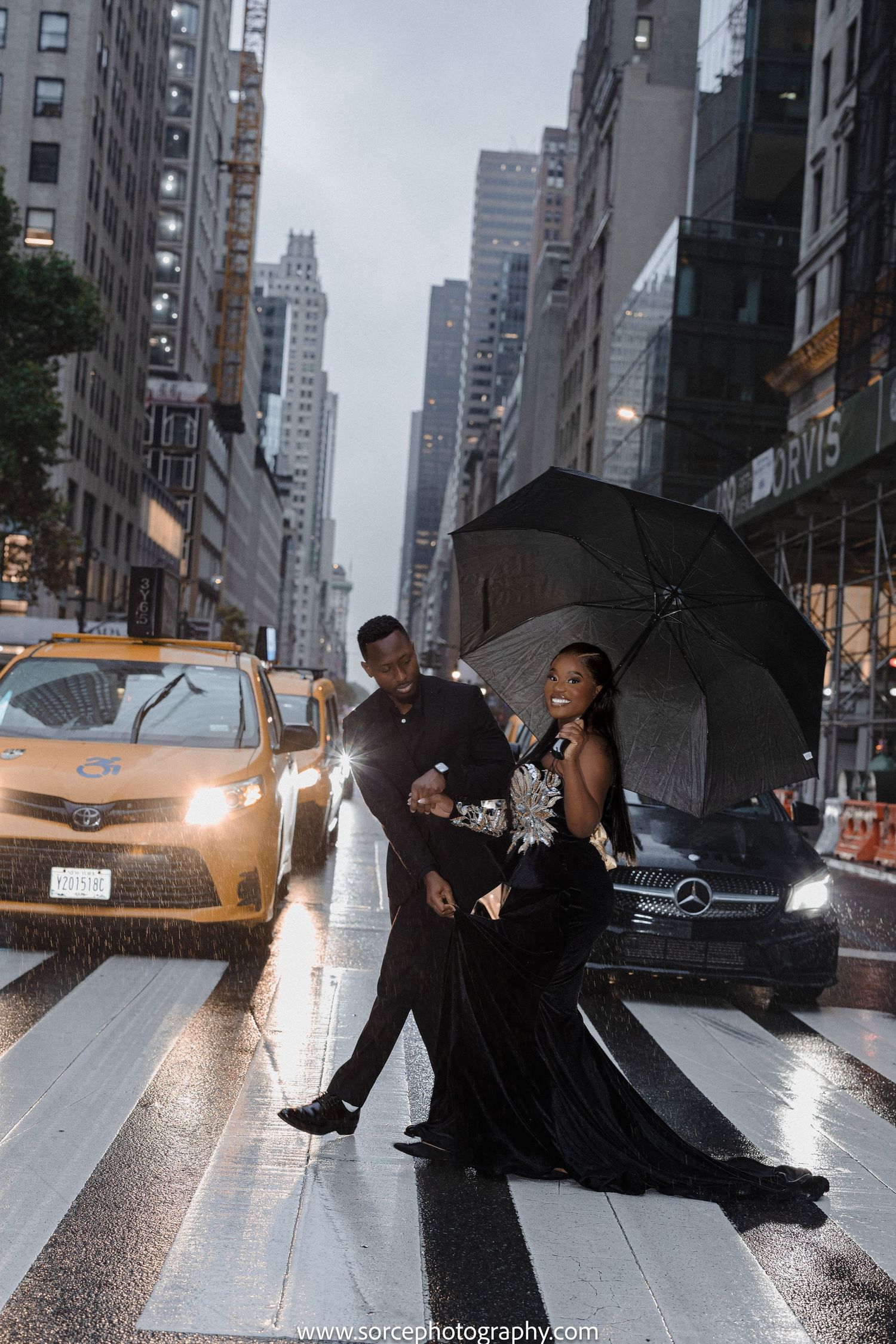 Couple in formal attire posing with umbrella on rainy city street with yellow taxi in background.