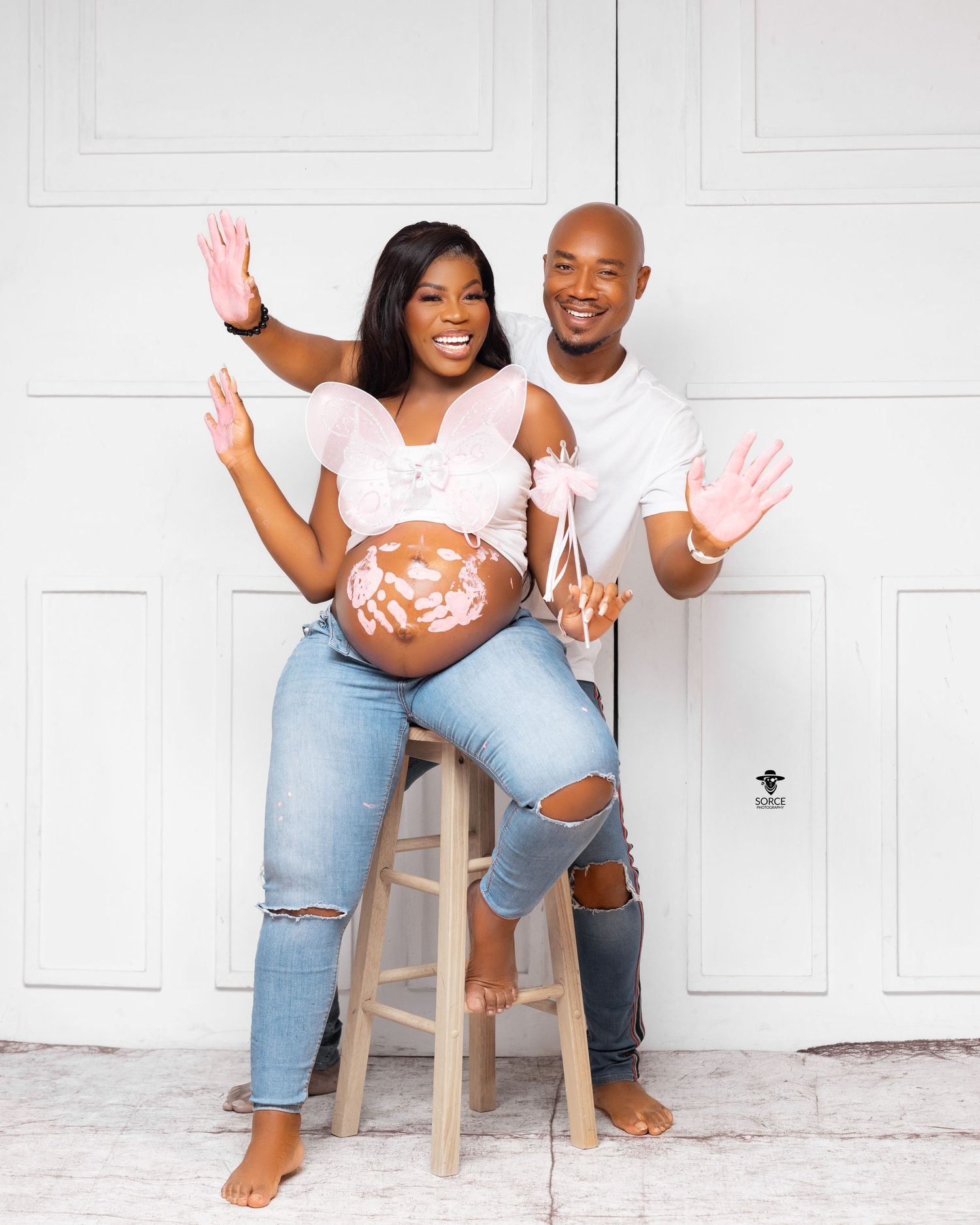 A couple in casual attire sitting on white wooden stools laughing and waving during a photoshoot.