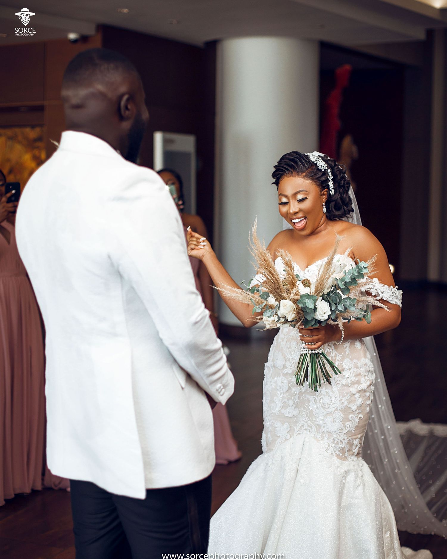 Joyful bride holding a beautiful white and green bouquet shares a happy first look moment in an indoor venue.