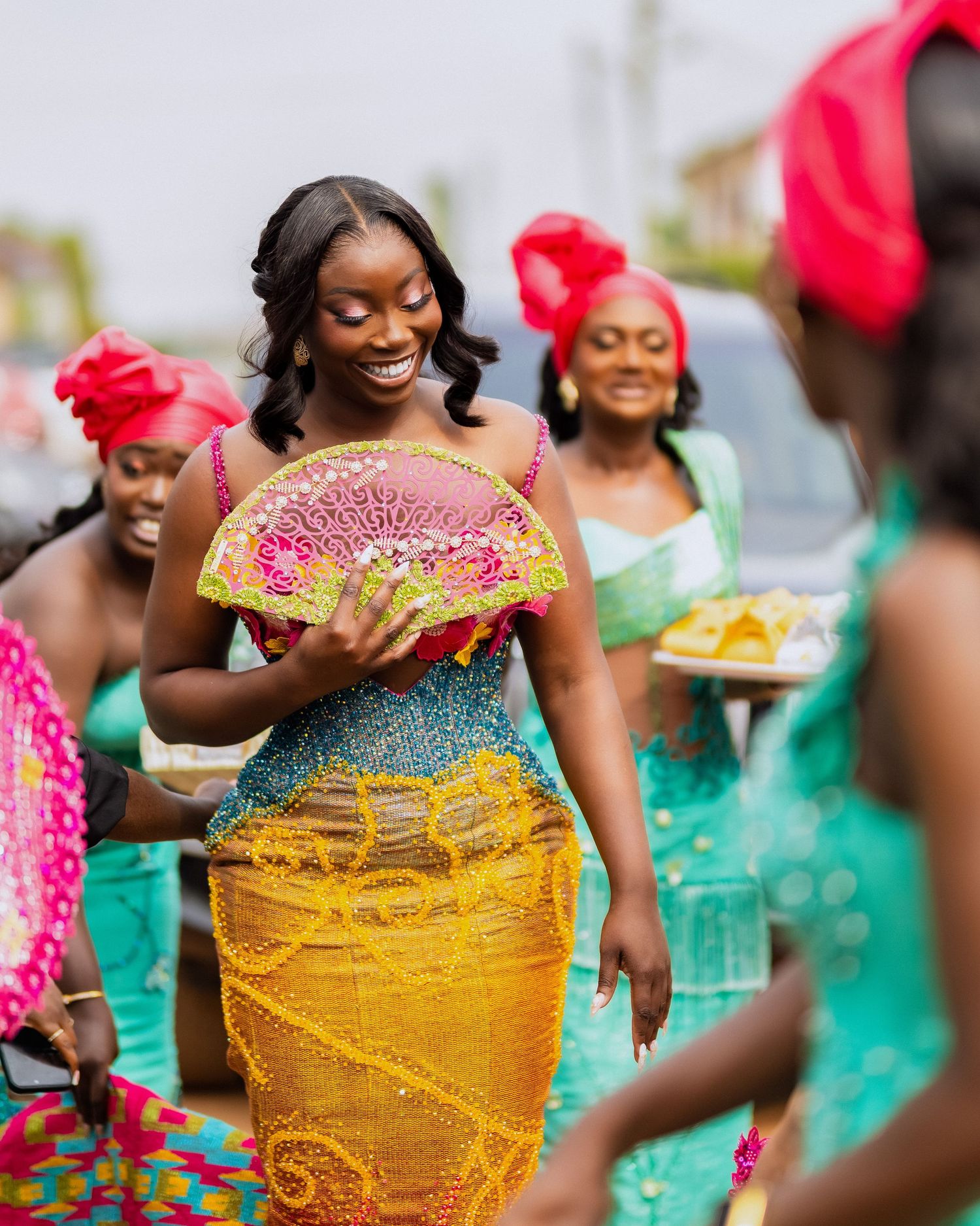 Women in vibrant traditional African attire celebrate at a cultural ceremony, with the lead woman holding a decorative fan.
