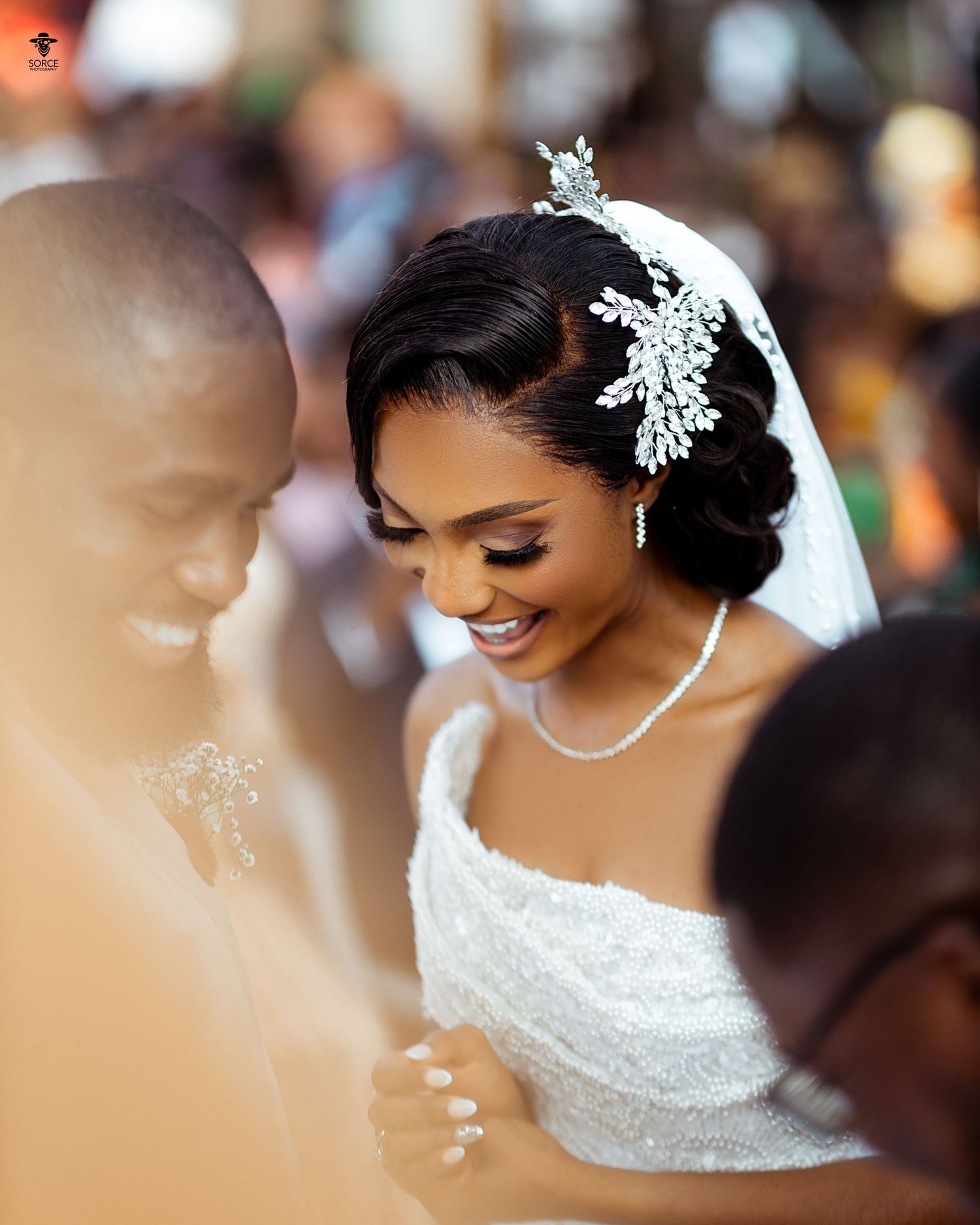 A radiant bride wearing an elegant lace gown, crystal hair accessory and pearl necklace smiles during her wedding ceremony.